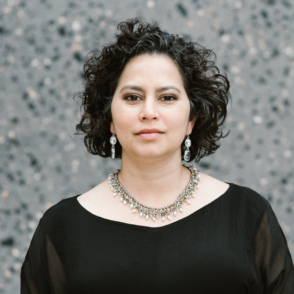 Woman with curly hair wearing earrings and a necklace, standing outdoors against a textured gray background.
