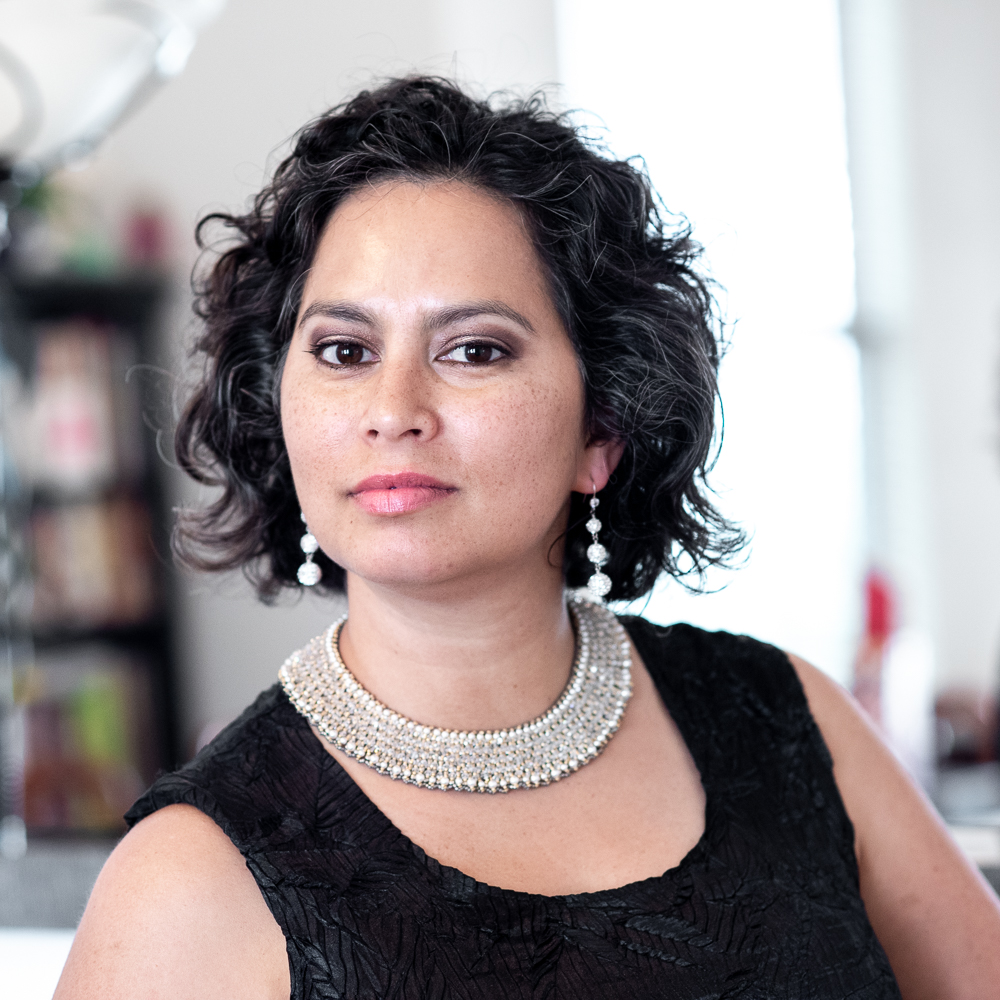 Woman with short curly hair wearing a black top, pearl necklace, and earrings, looking at the camera in a bright room.
