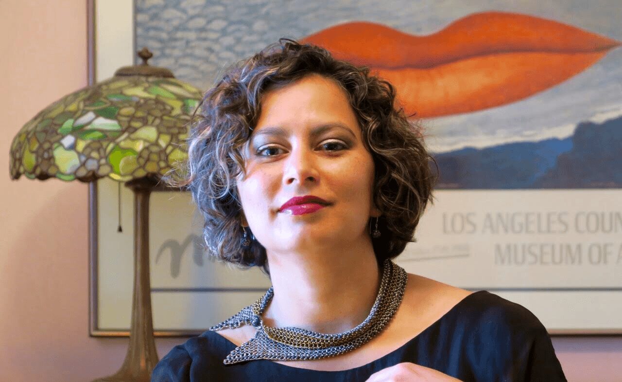 Woman with short curly hair wearing a necklace, standing indoors with a lamp and artwork in the background.