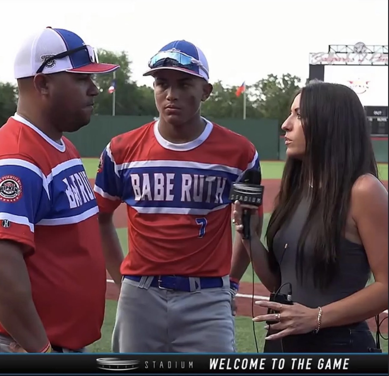 Three people standing on a baseball field, two men in red and blue uniforms and a woman with a microphone, during an interview.