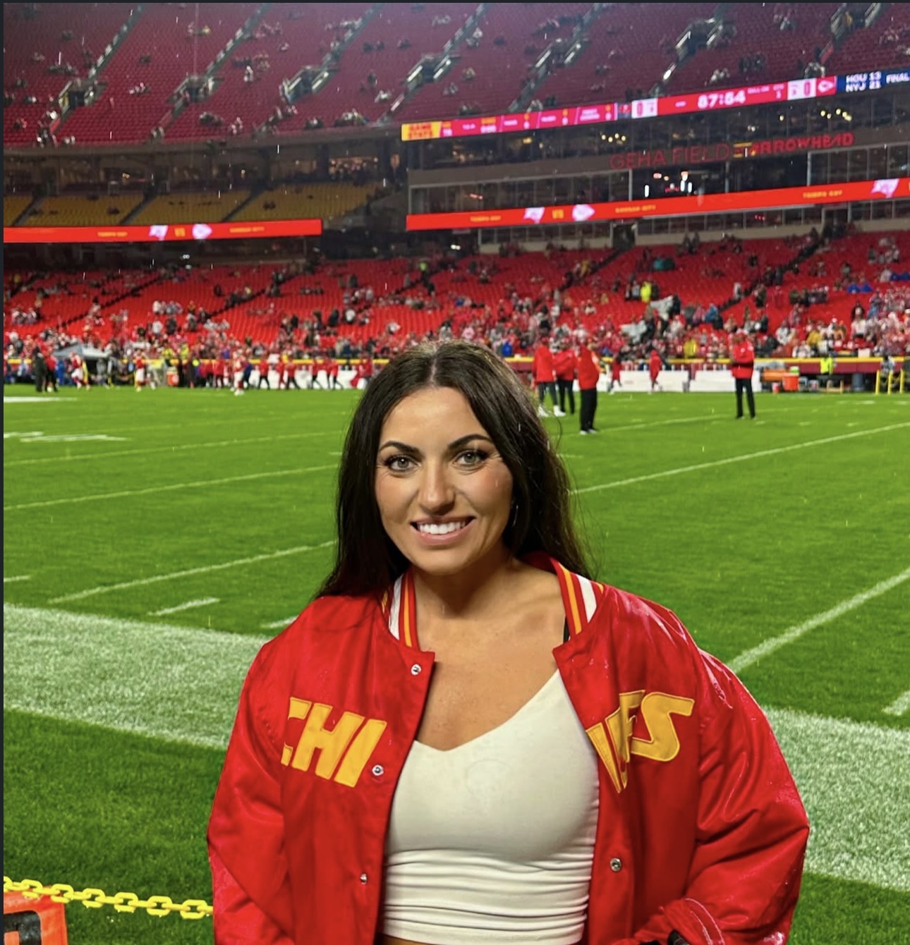 Woman smiling on a football field wearing a red jacket, stadium seats in background