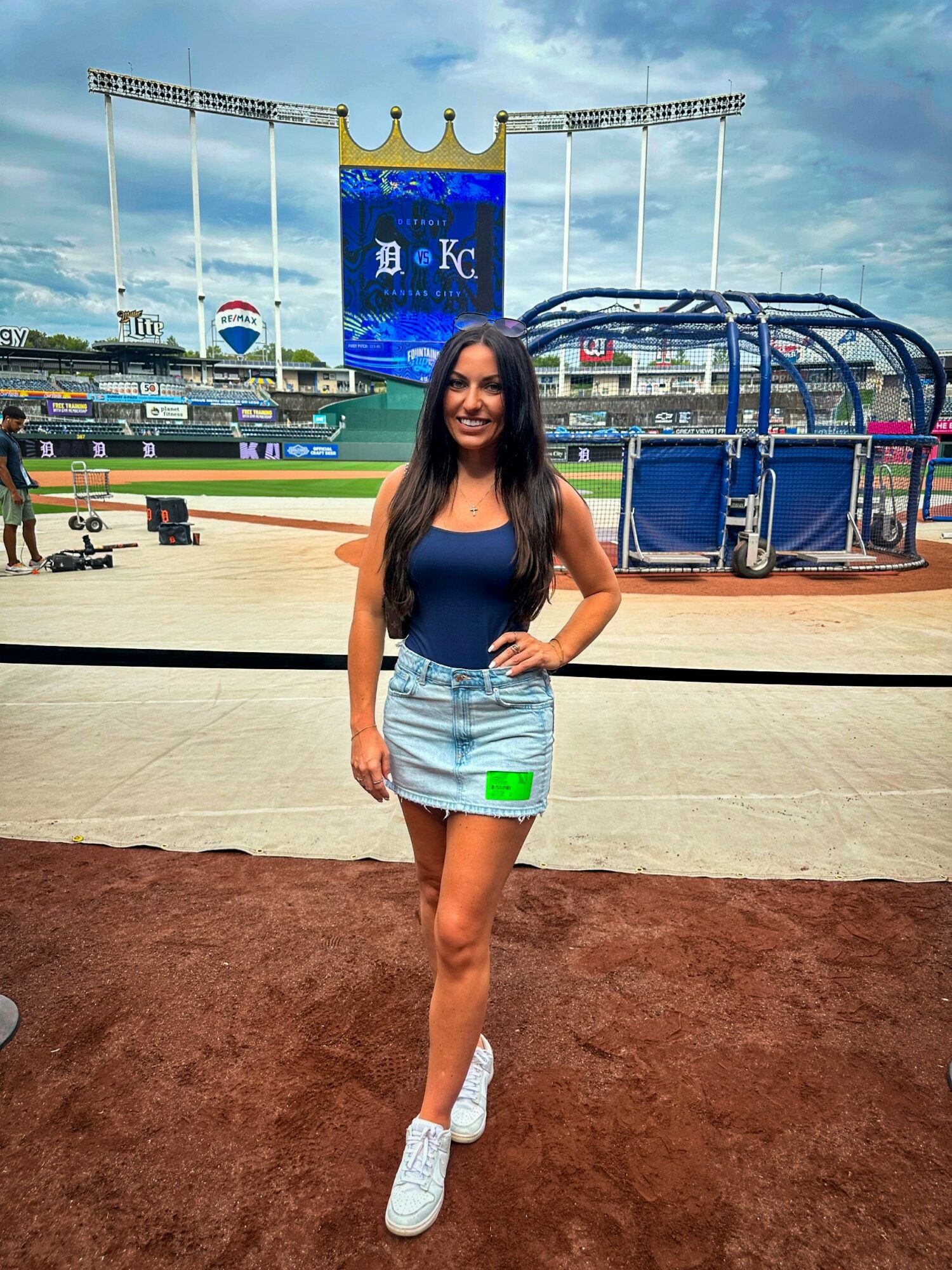 Young woman standing on baseball field with stadium and scoreboard in background, smiling, wearing a blue top and denim skirt.