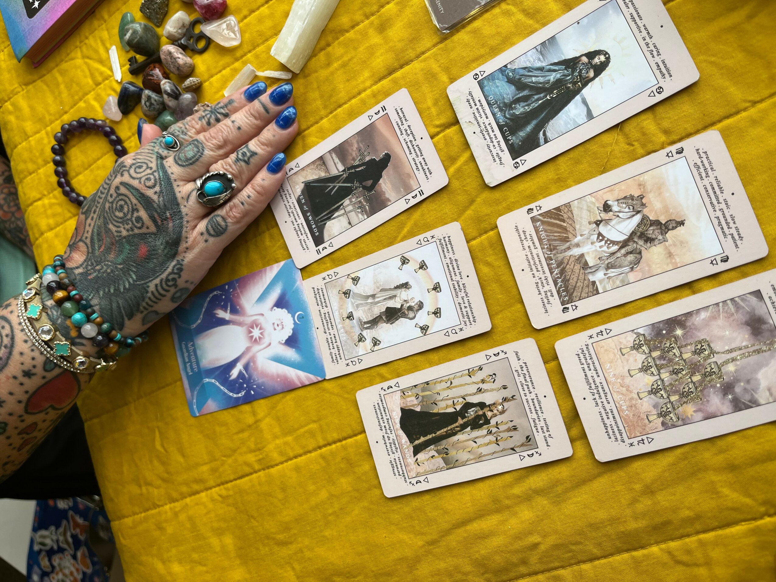 Hand with tattoos and jewelry over tarot cards spread on yellow fabric, with crystals nearby.