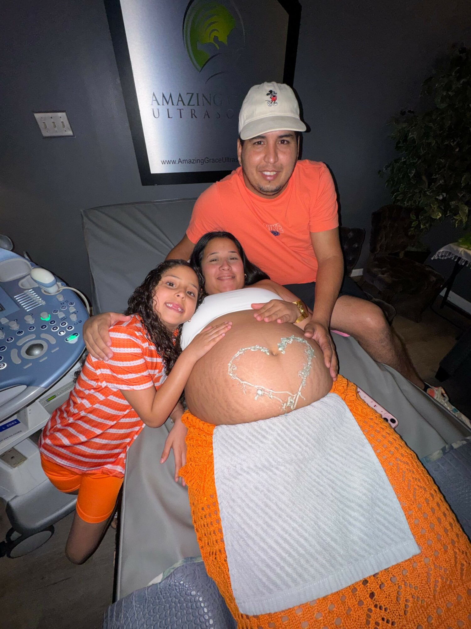 Pregnant woman lying on a medical bed with a woman and a girl, all smiling, in a clinical setting.
