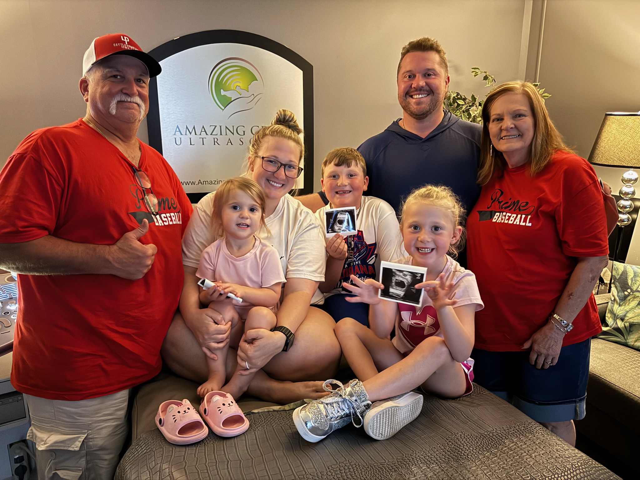 Group of seven people, including children, smiling indoors with a sign in the background, some holding photos.