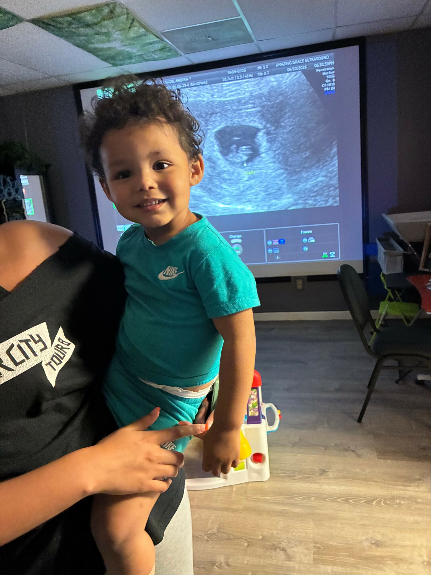 Child with curly hair in a turquoise shirt interacts with a person, in a room with a large screen displaying an ultrasound image.