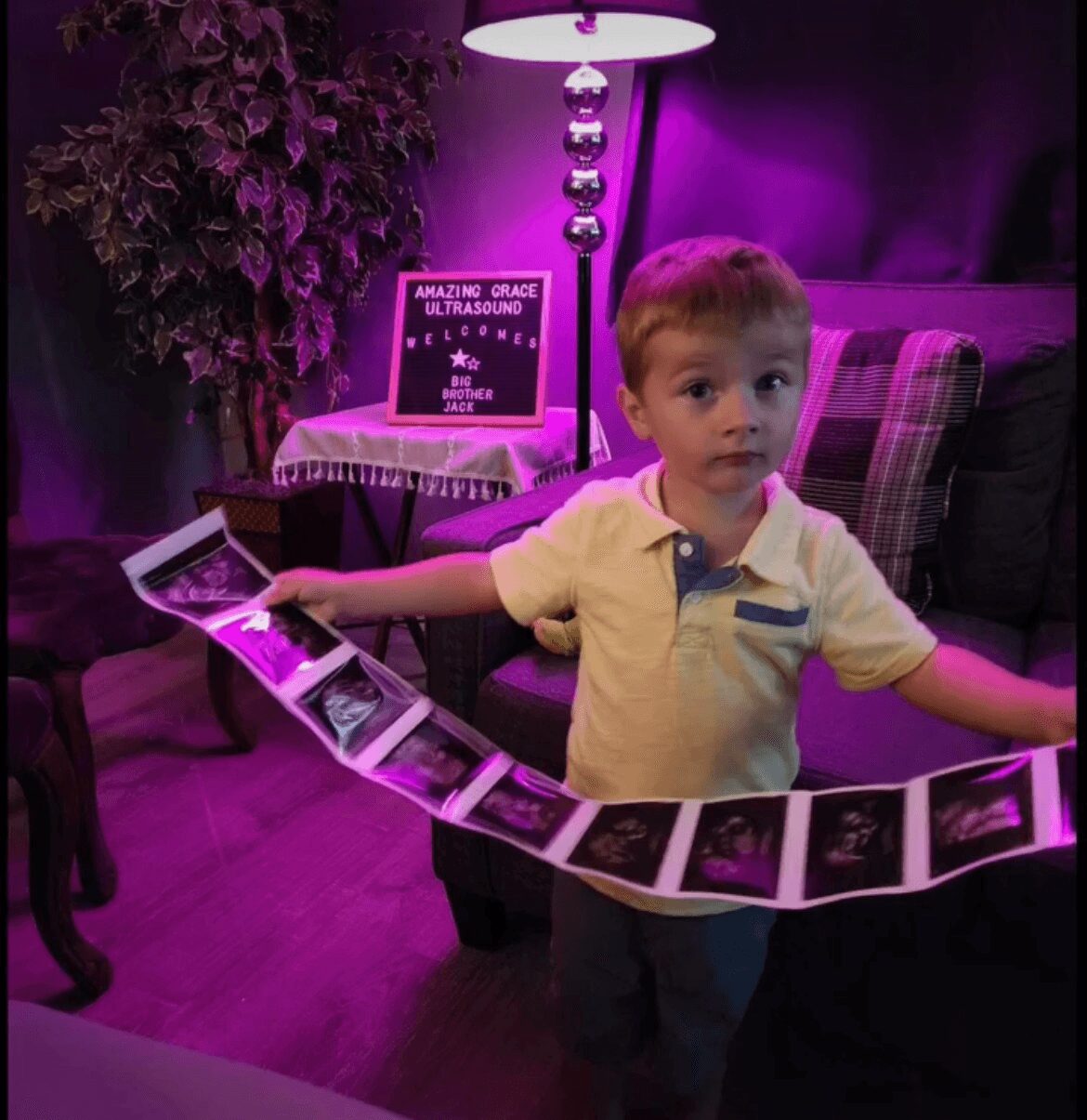 Child holding a string of ultrasound images, standing in a room with purple lighting and a sign about ultrasound, with a lamp and plants in the background.