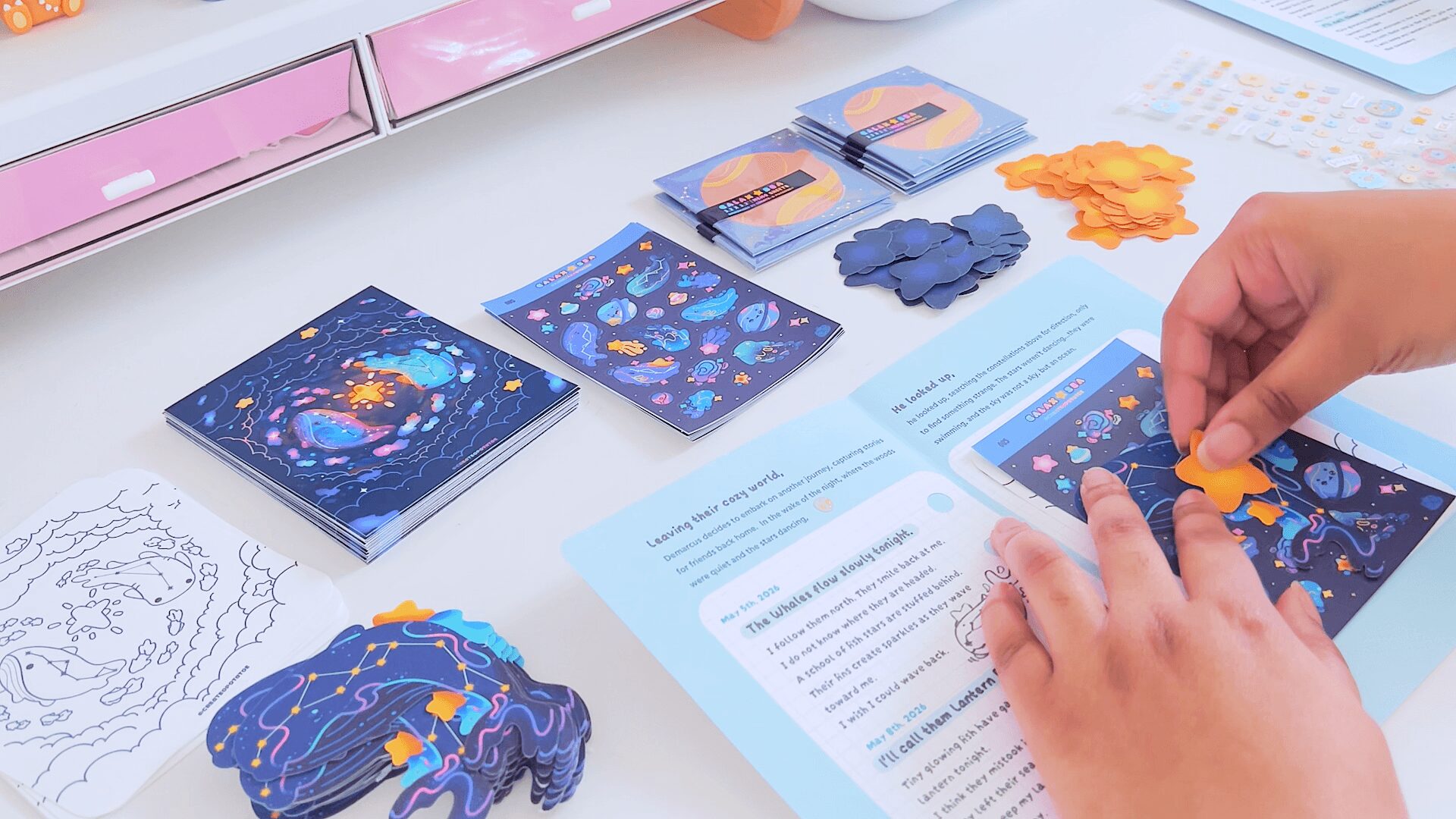 Person's hands holding a booklet with space-themed illustrations, surrounded by space cards, clouds, and other educational materials on a table.