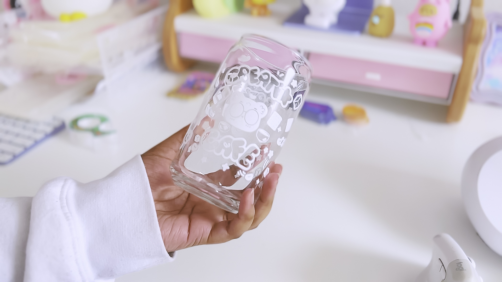 Person holding a clear glass with white decorative patterns, background shows colorful objects and a pink shelf, workspace setting