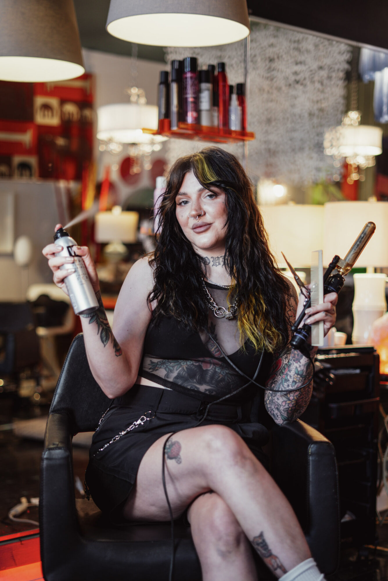Woman with dark hair and tattoos sitting in a salon chair holding a spray bottle and a curling iron, with salon equipment in background.