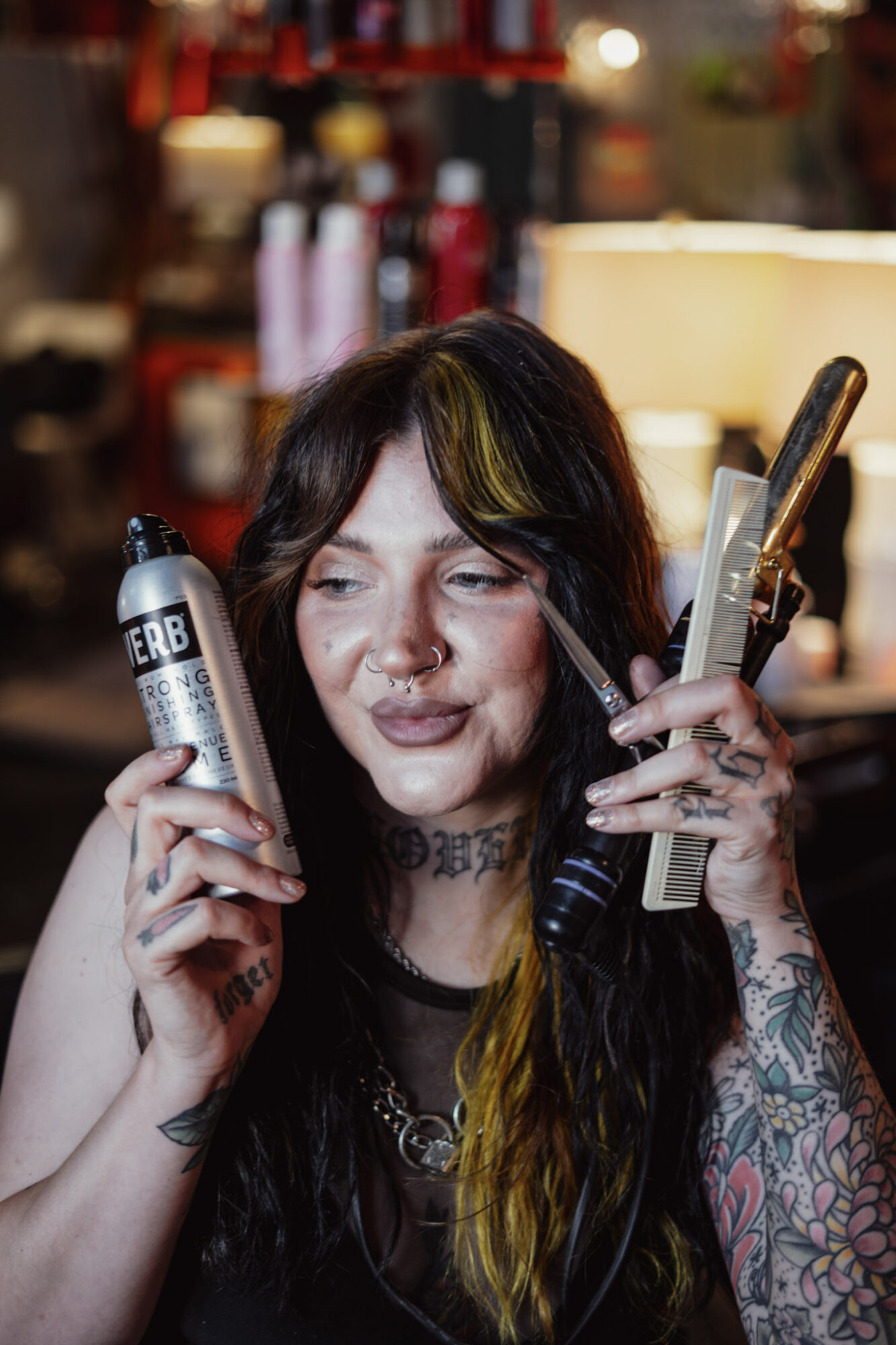 Woman with dark hair and tattoos holding a spray can and a phone in a salon or barbershop.