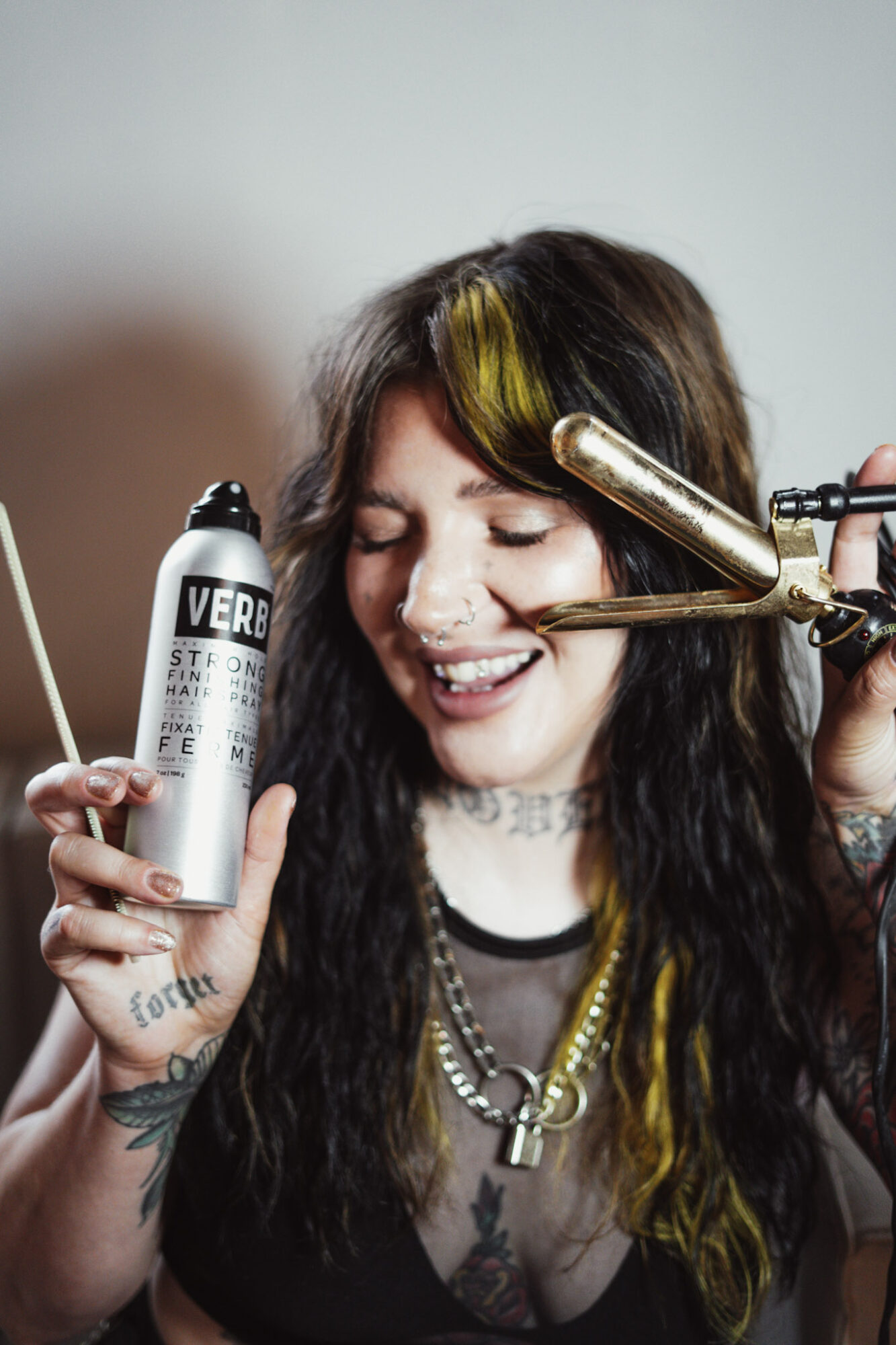 Woman with dark hair and yellow streaks, smiling, holding a can of hair spray, with a curling iron on her hair.