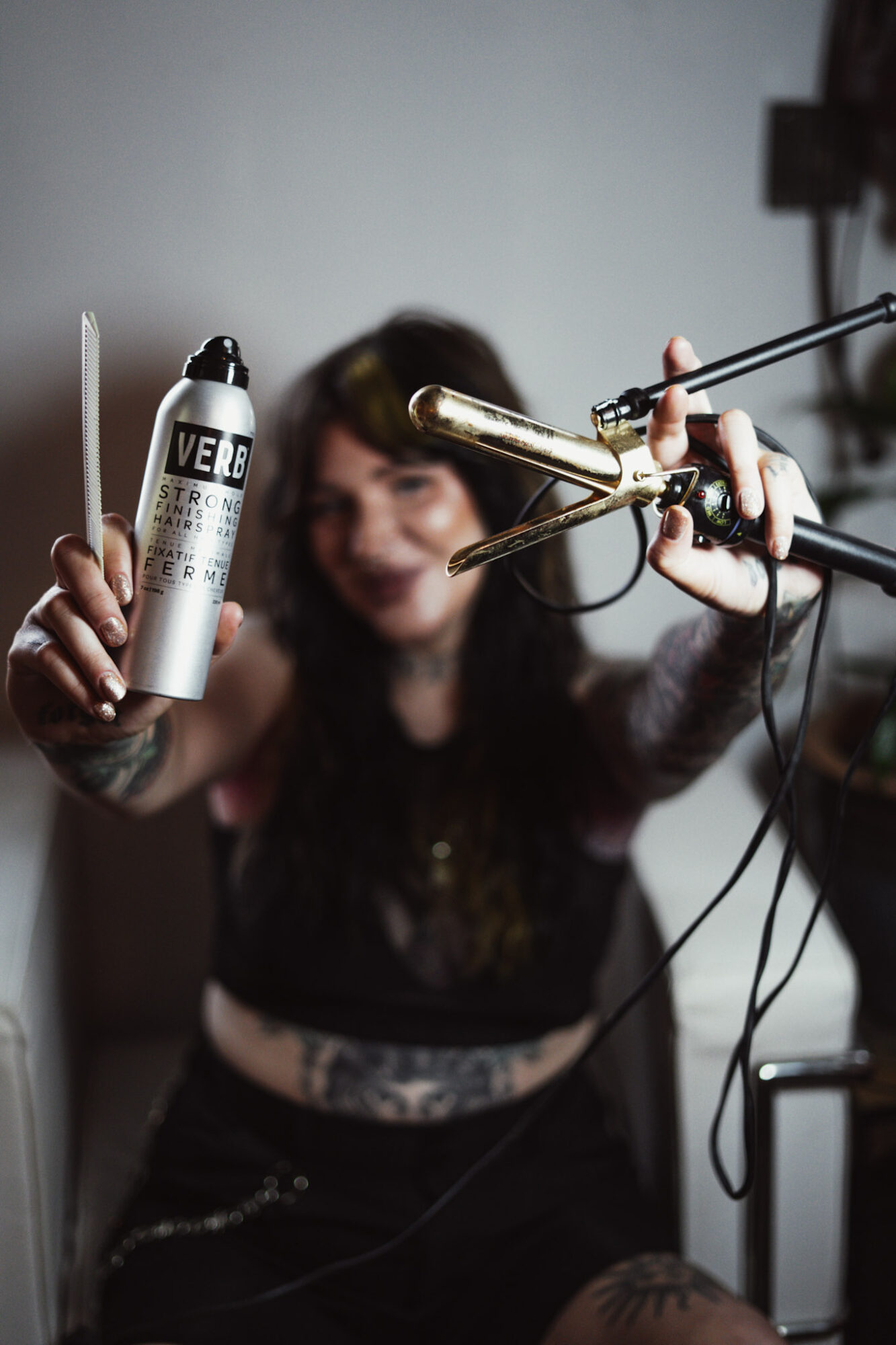 Person holding a can of hairspray and a curling iron, smiling, with a blurred background.