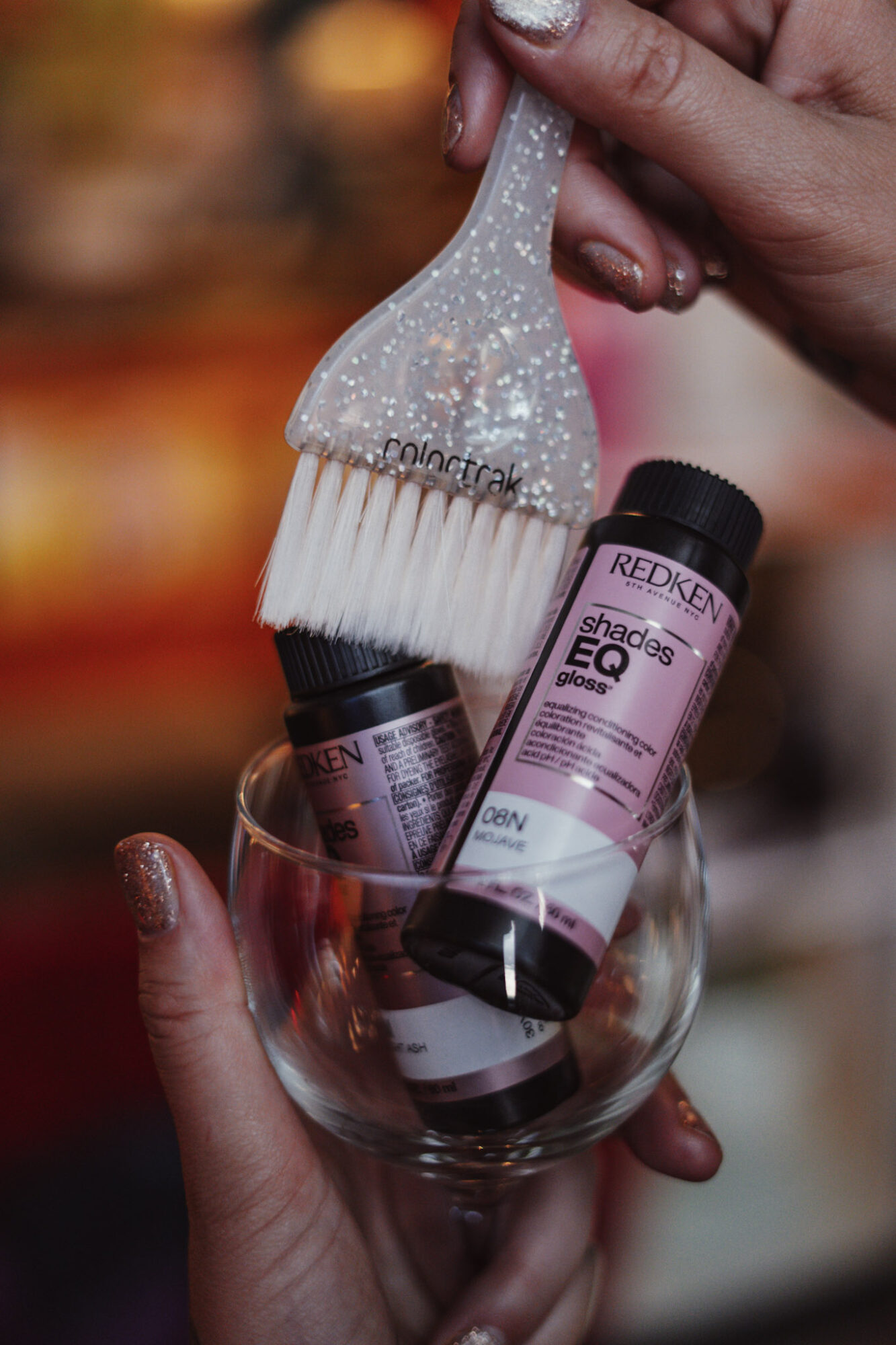 Person holding a glass with skincare bottles and a brush, with a blurred background.