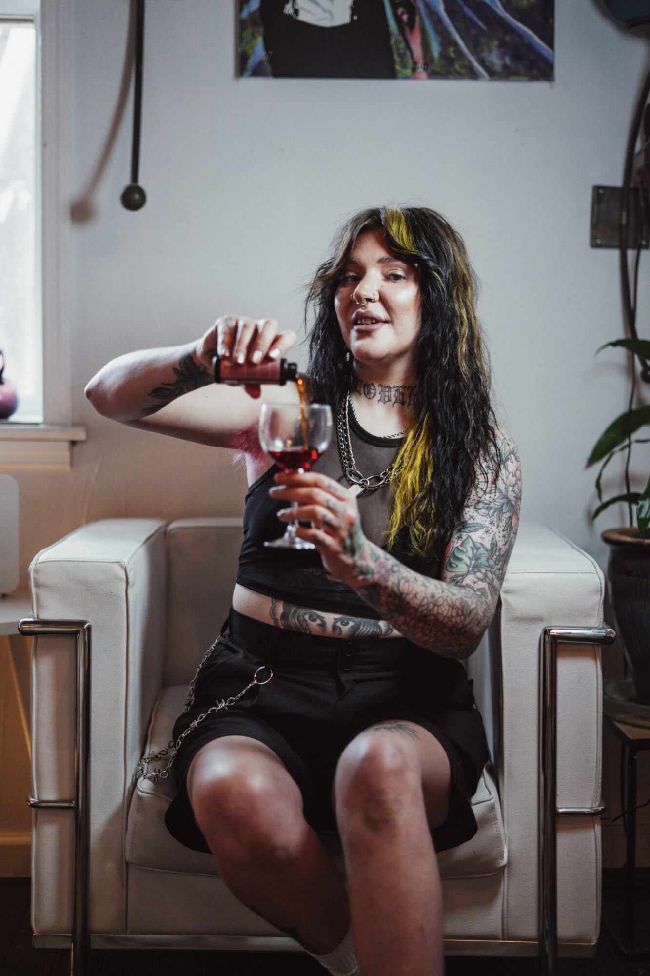 Young woman with long dark hair and tattoos, sitting on a sofa, pouring red wine into a glass, smiling.