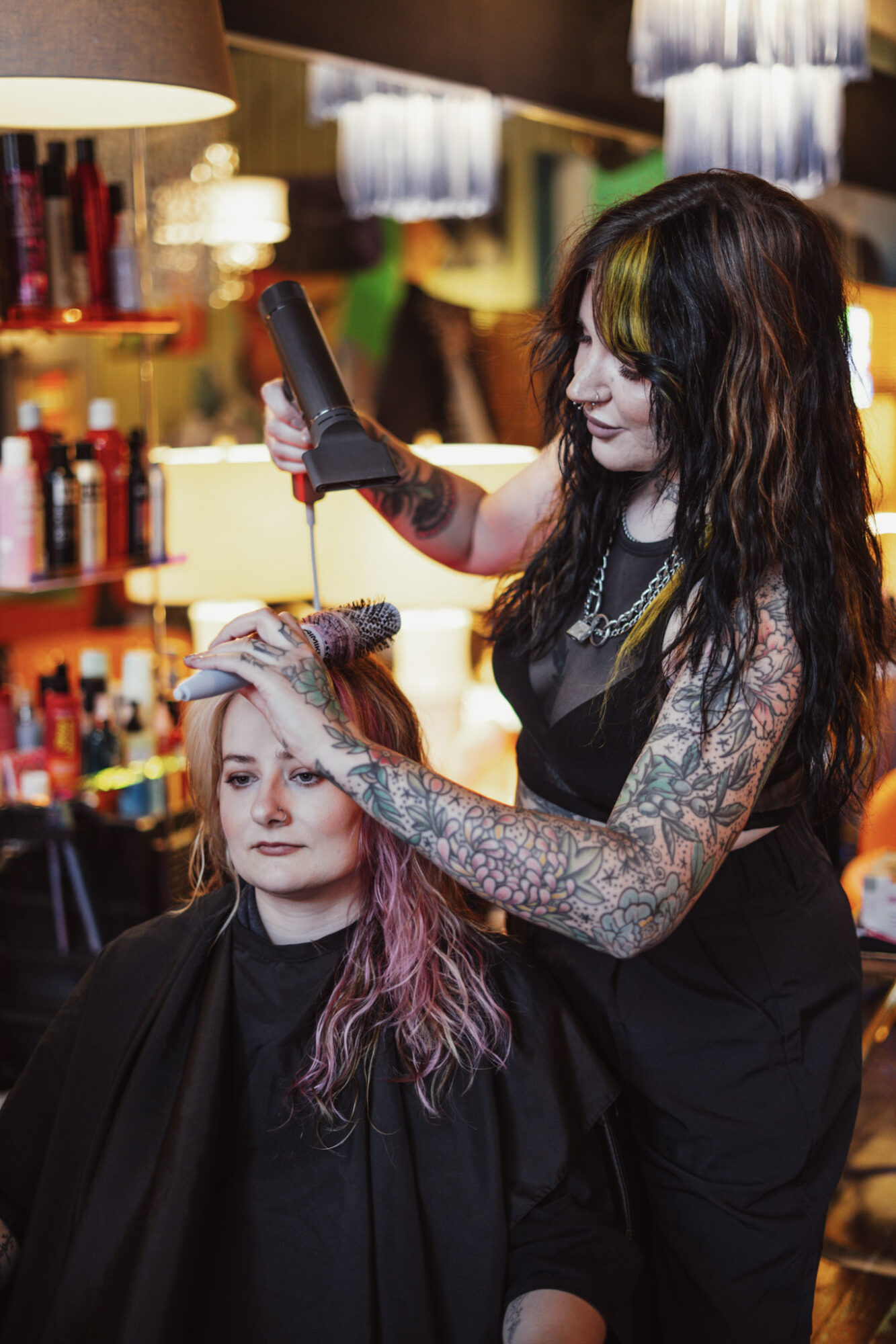 Hairdresser styles a woman's hair in a salon with colorful lighting and shelves of hair products, woman has pink and purple hair.
