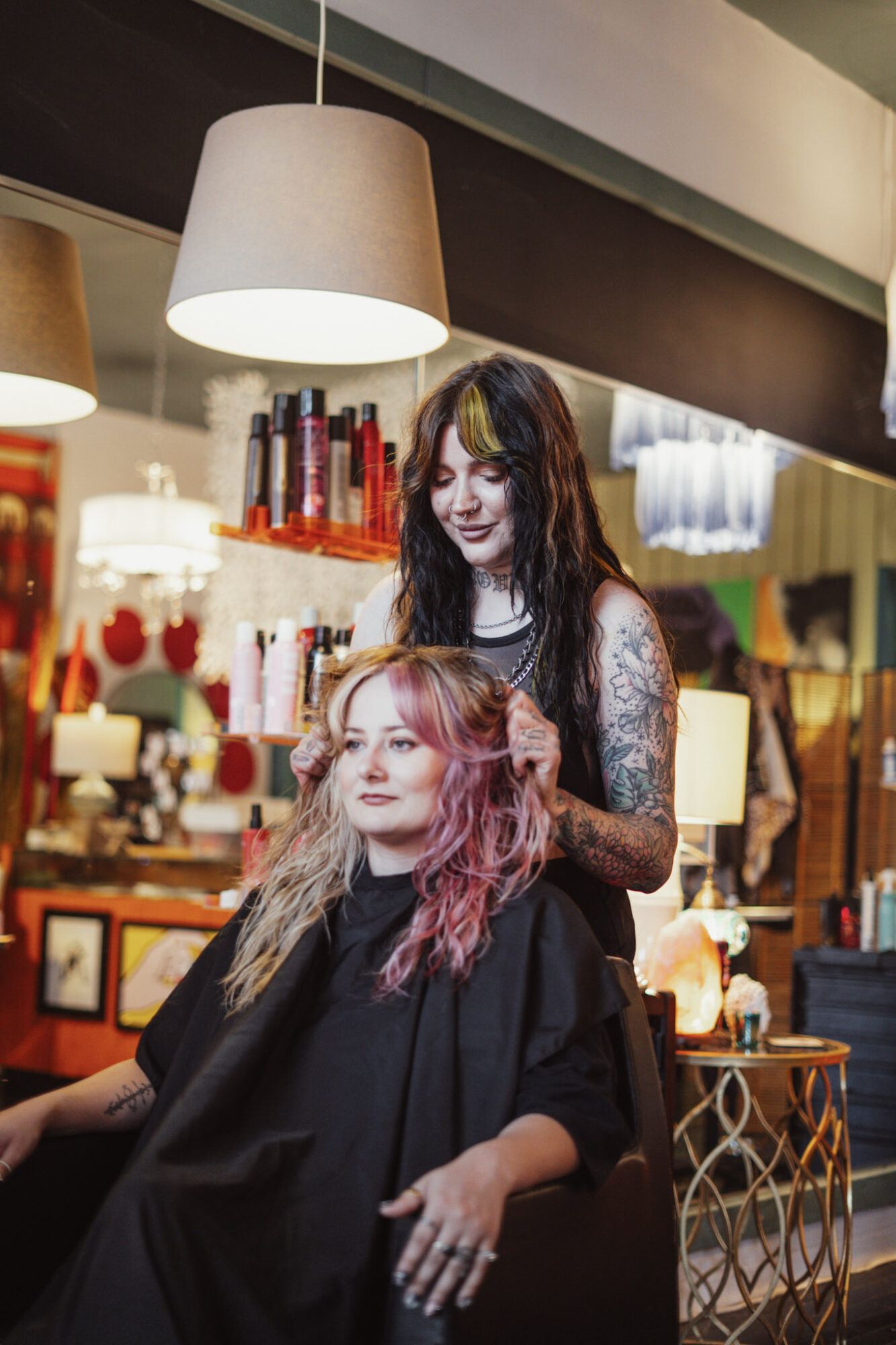 A hairstylist with dark hair and tattoos styles a woman's pink, wavy hair in a salon interior.