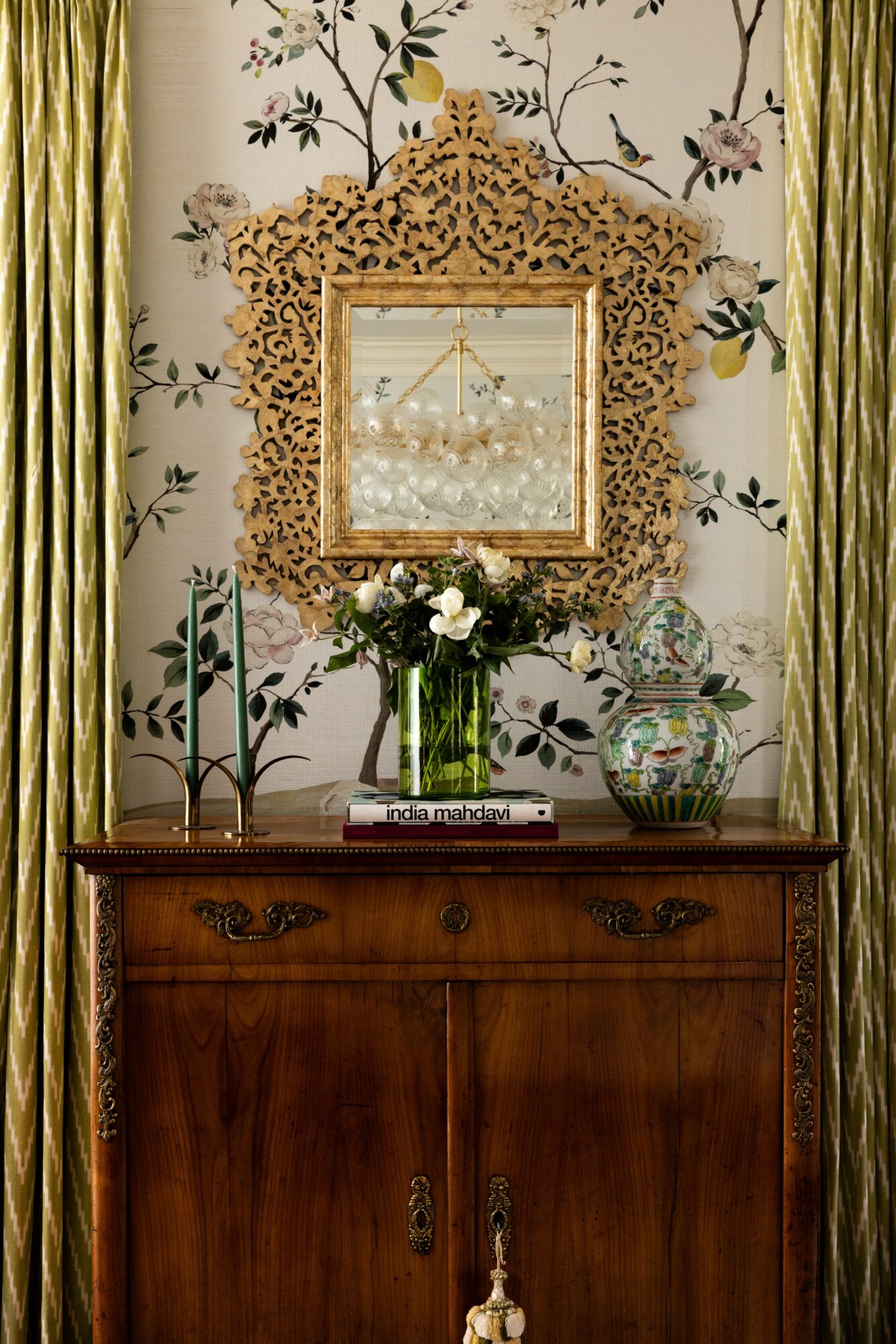 Decorative mirror with intricate gold frame above a wooden sideboard, with a vase of white roses and a glass fishbowl, against floral wallpaper.