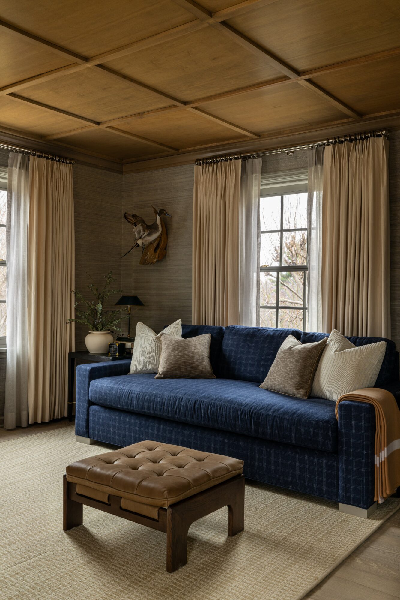 Living room with a blue sofa, beige pillows, wooden ceiling, and large windows with curtains.
