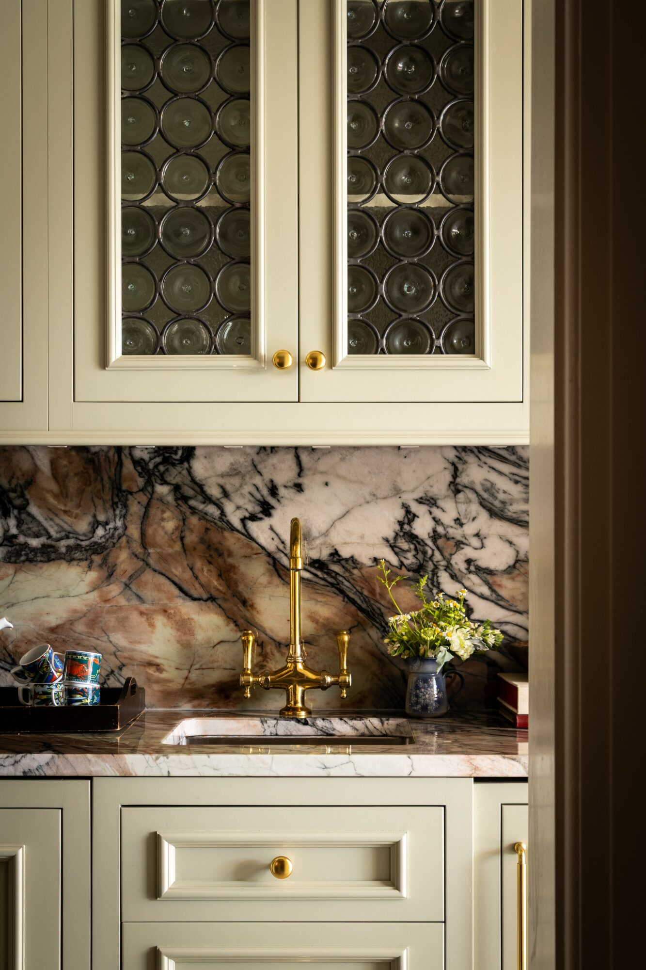 Kitchen cabinet with glass doors, marble countertop, gold faucet, and a small plant in a vase.