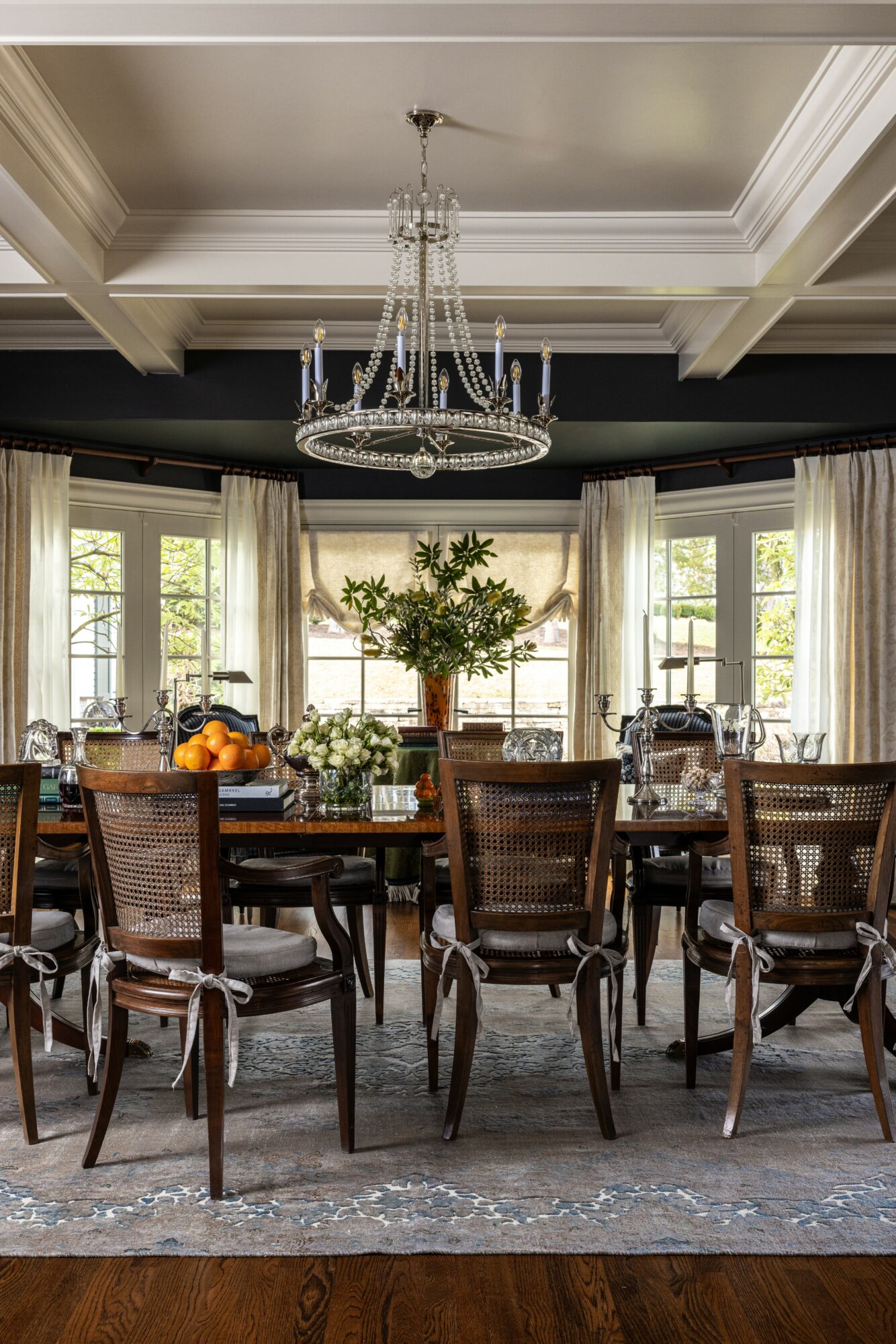 Elegant dining room with a large wooden table, chairs, a chandelier, and a window with curtains, natural light, and a plant centerpiece.