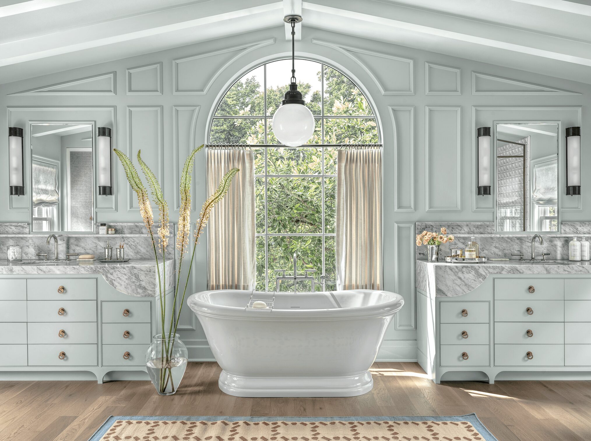 Elegant bathroom with a bathtub centered in front of a large arched window, flanked by two vanities with mirrors, in a bright room.