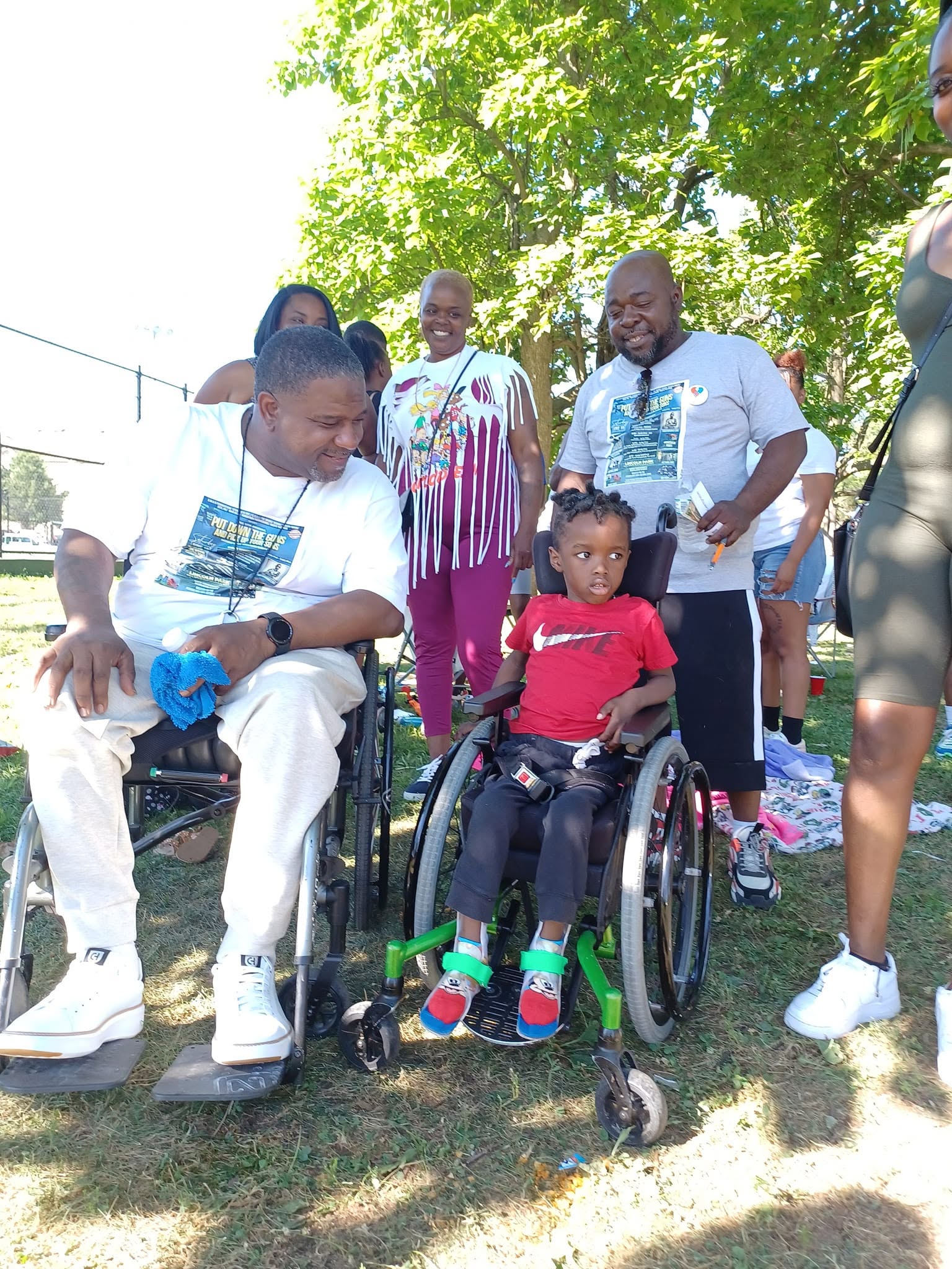 Group of people outdoors, some standing and one person in a wheelchair, under green trees, sunny day.