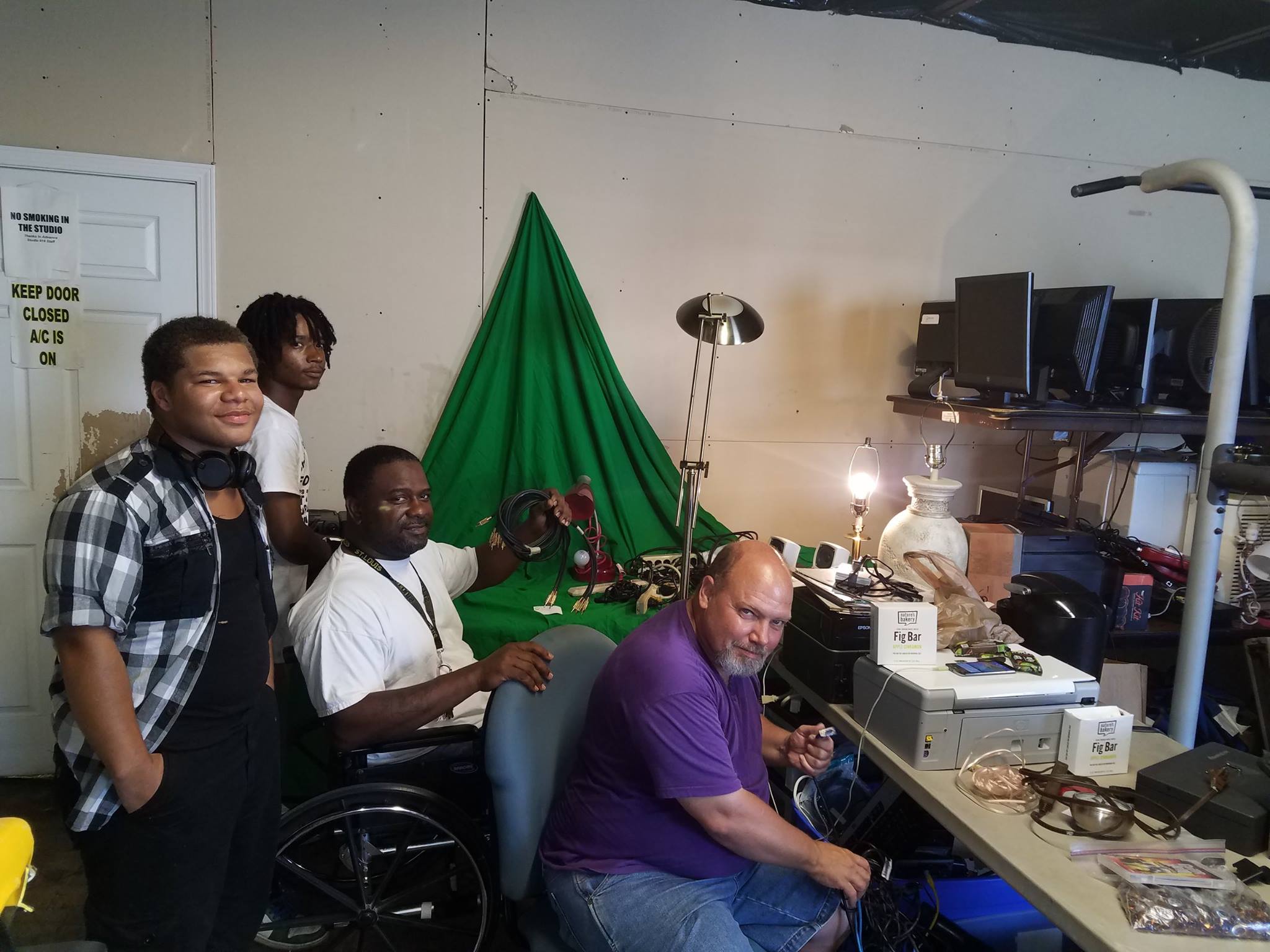 Group of five people in a room with a green backdrop, some sitting at a desk with electronic equipment, one standing.