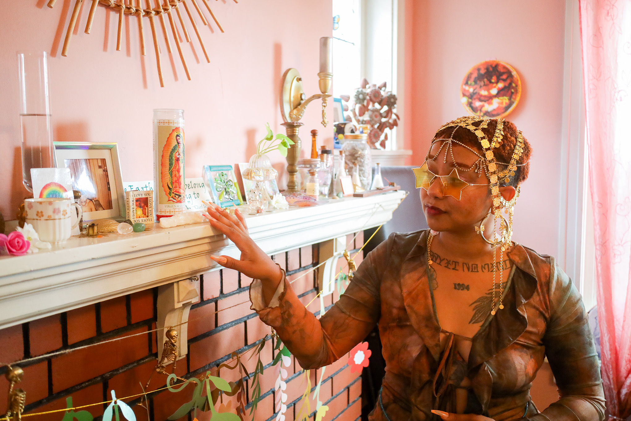 Woman with elaborate jewelry and tattoos reaches toward a decorated fireplace mantel in a pink room.