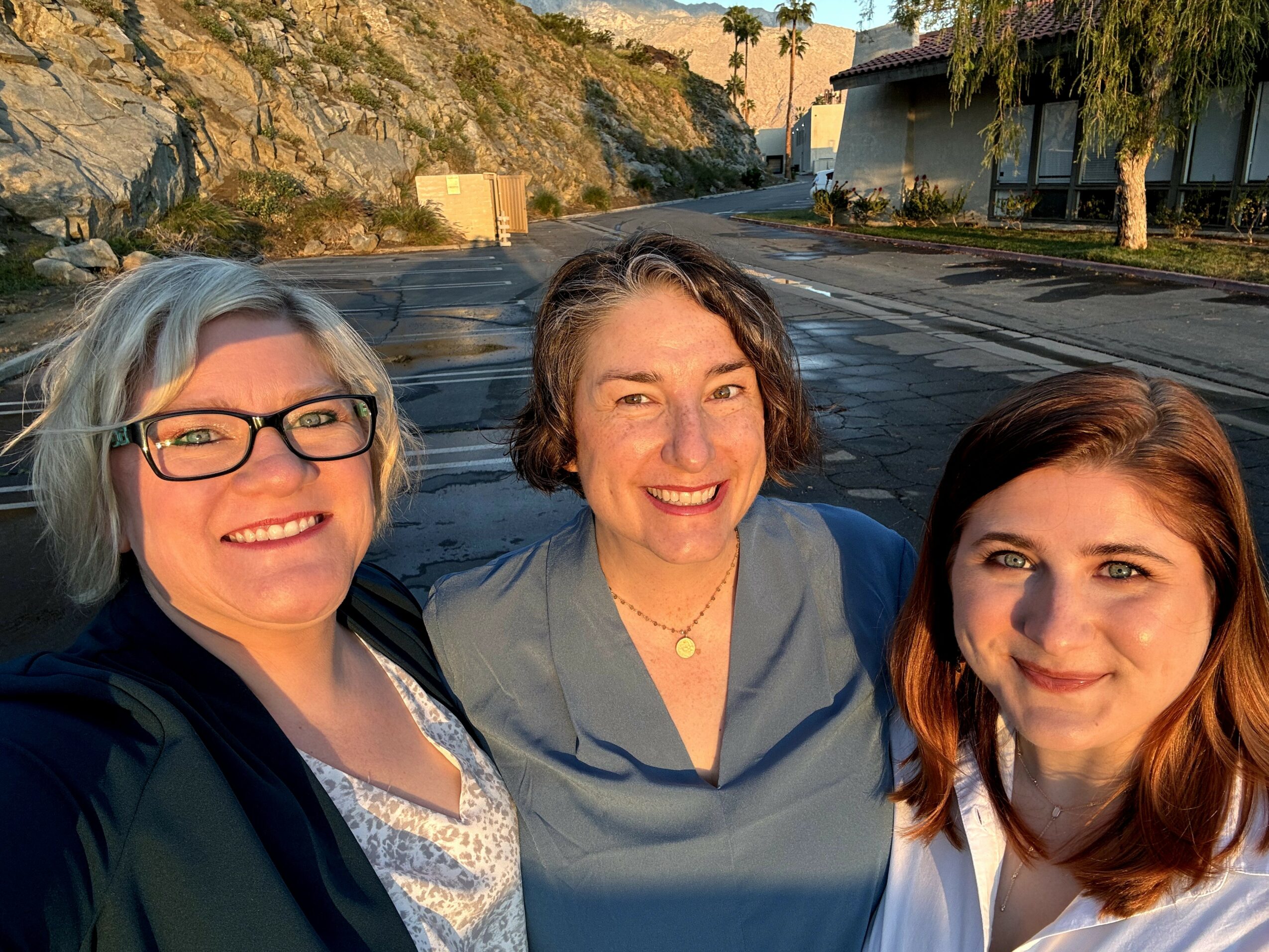 Three women smiling outdoors on a sunny day, with trees and buildings in the background.
