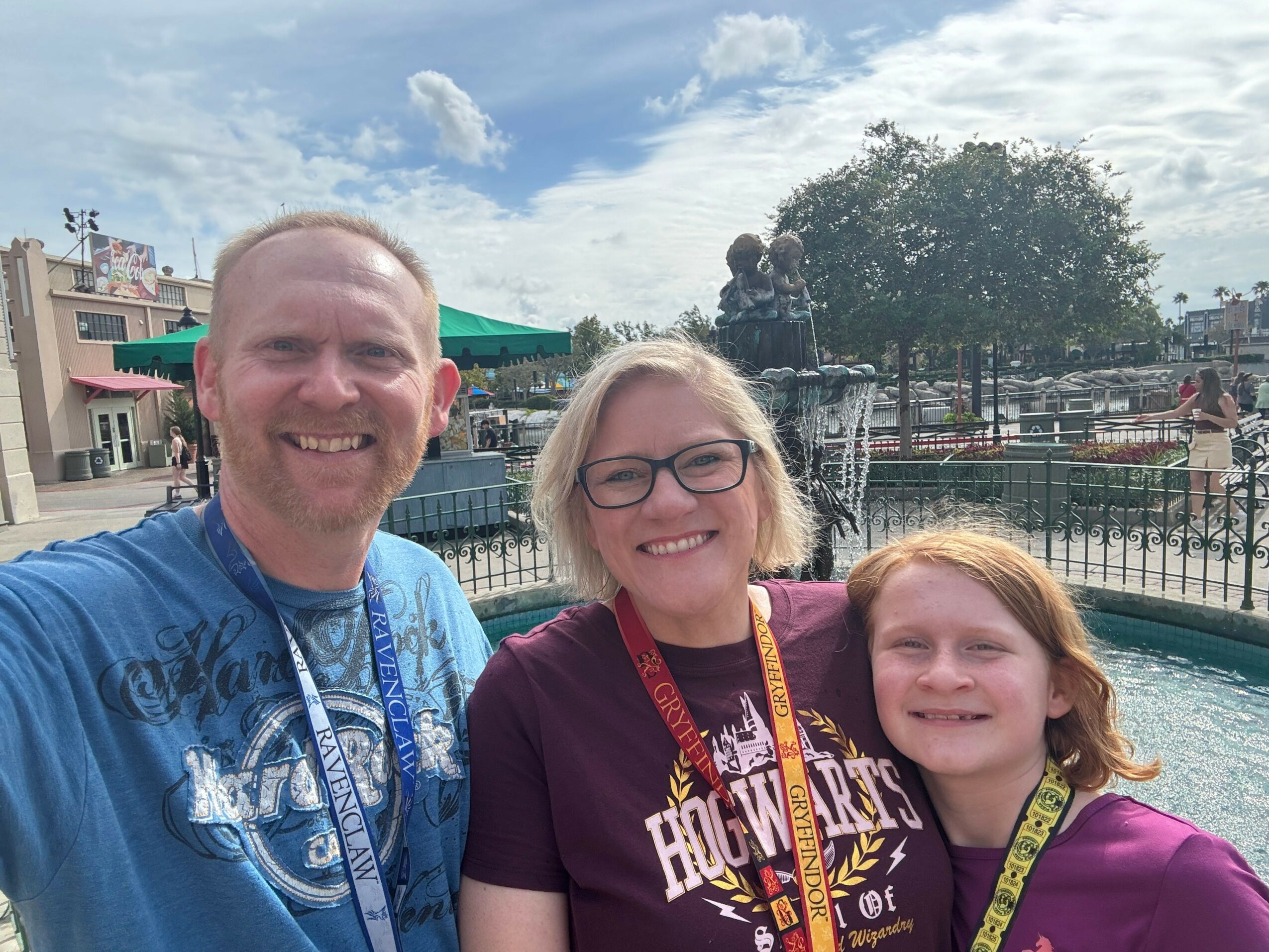 Three people smiling outdoors with a fence, trees, and cloudy sky in the background.