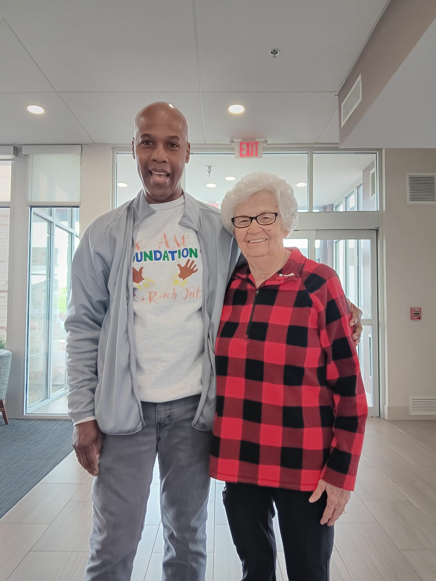 Two people standing indoors, one with glasses and a red checkered jacket, the other with a white shirt and gray jacket.