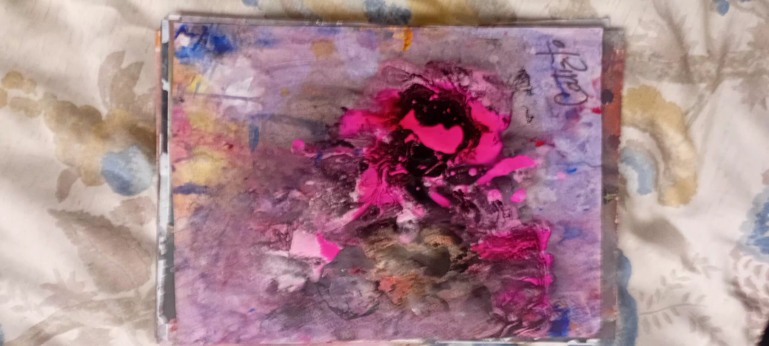 Abstract painting with pink, purple, and blue colors on a rectangular canvas, placed on a patterned fabric surface.
