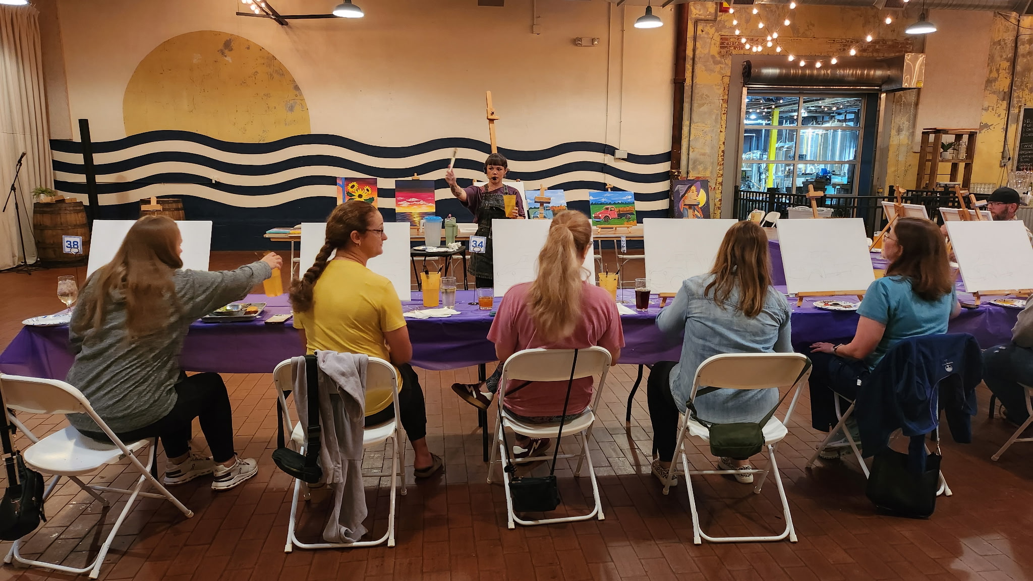 People seated at a long table in a decorated indoor space, with a mural and string lights in the background.