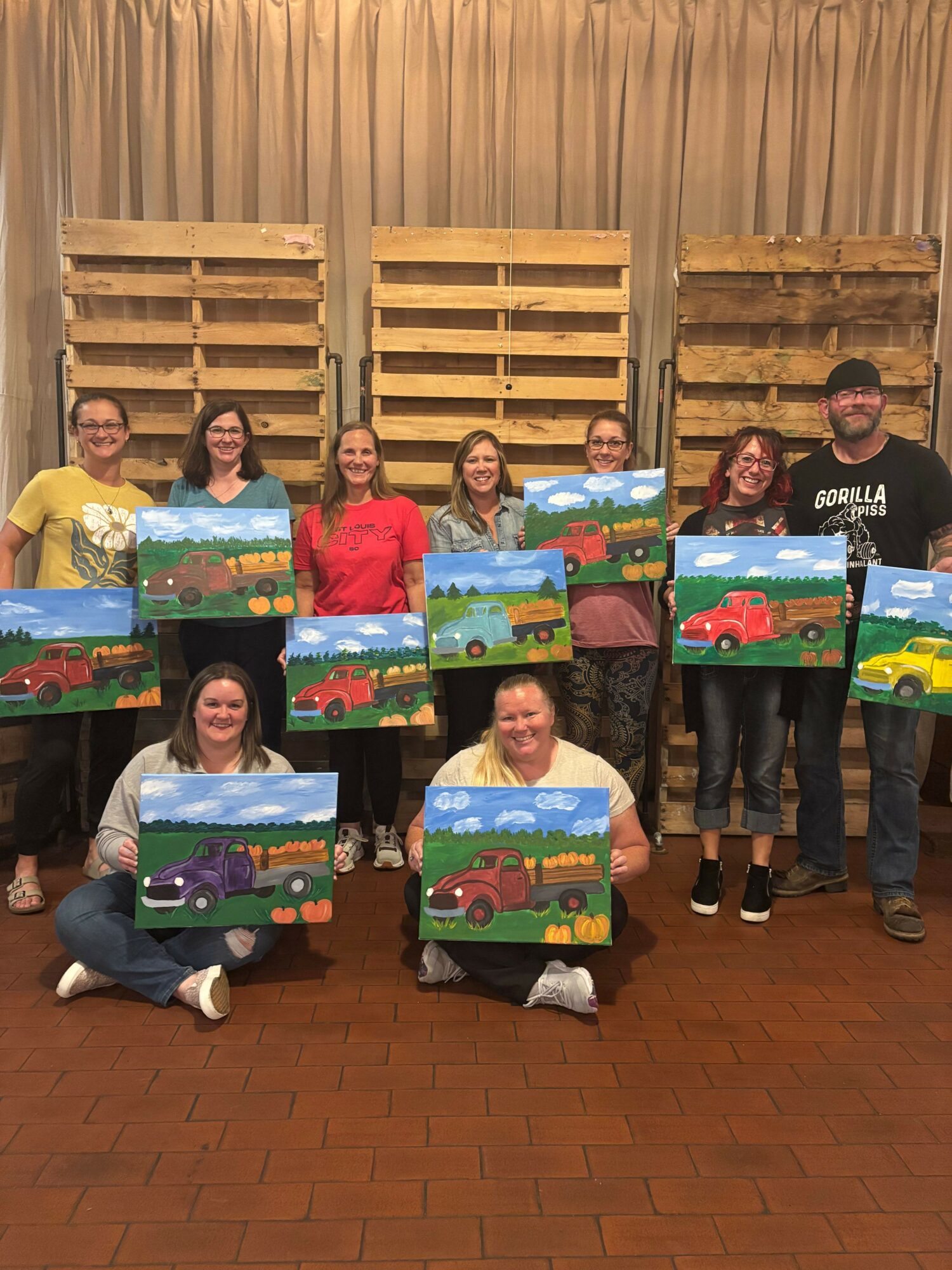 Group of ten people holding colorful paintings of trucks and landscapes, standing in front of wooden pallets and curtains.