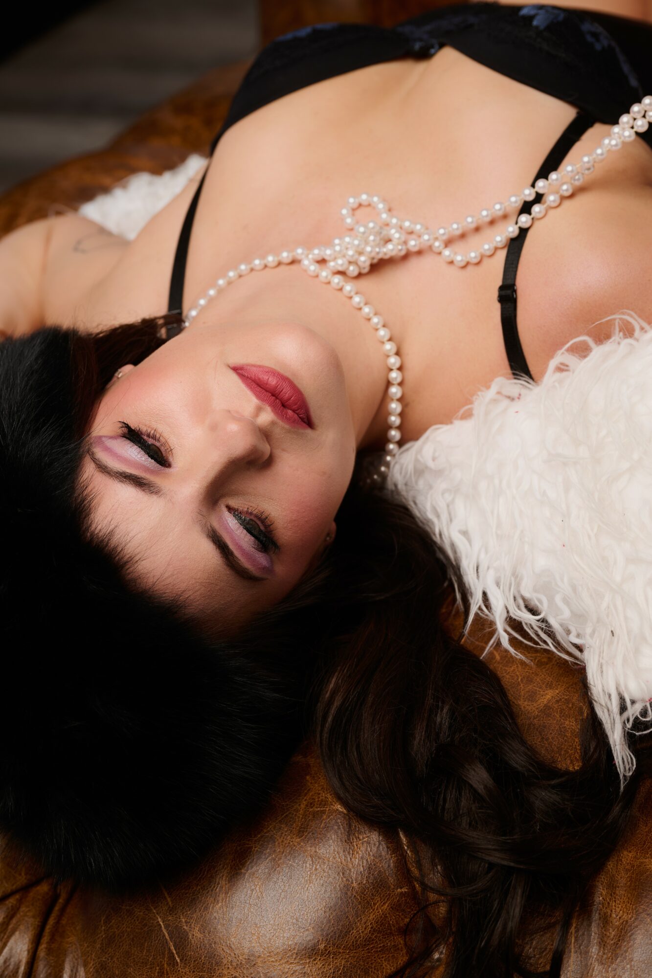 Woman lying down with dark hair, wearing pearl necklace and black lingerie, on a furry surface.
