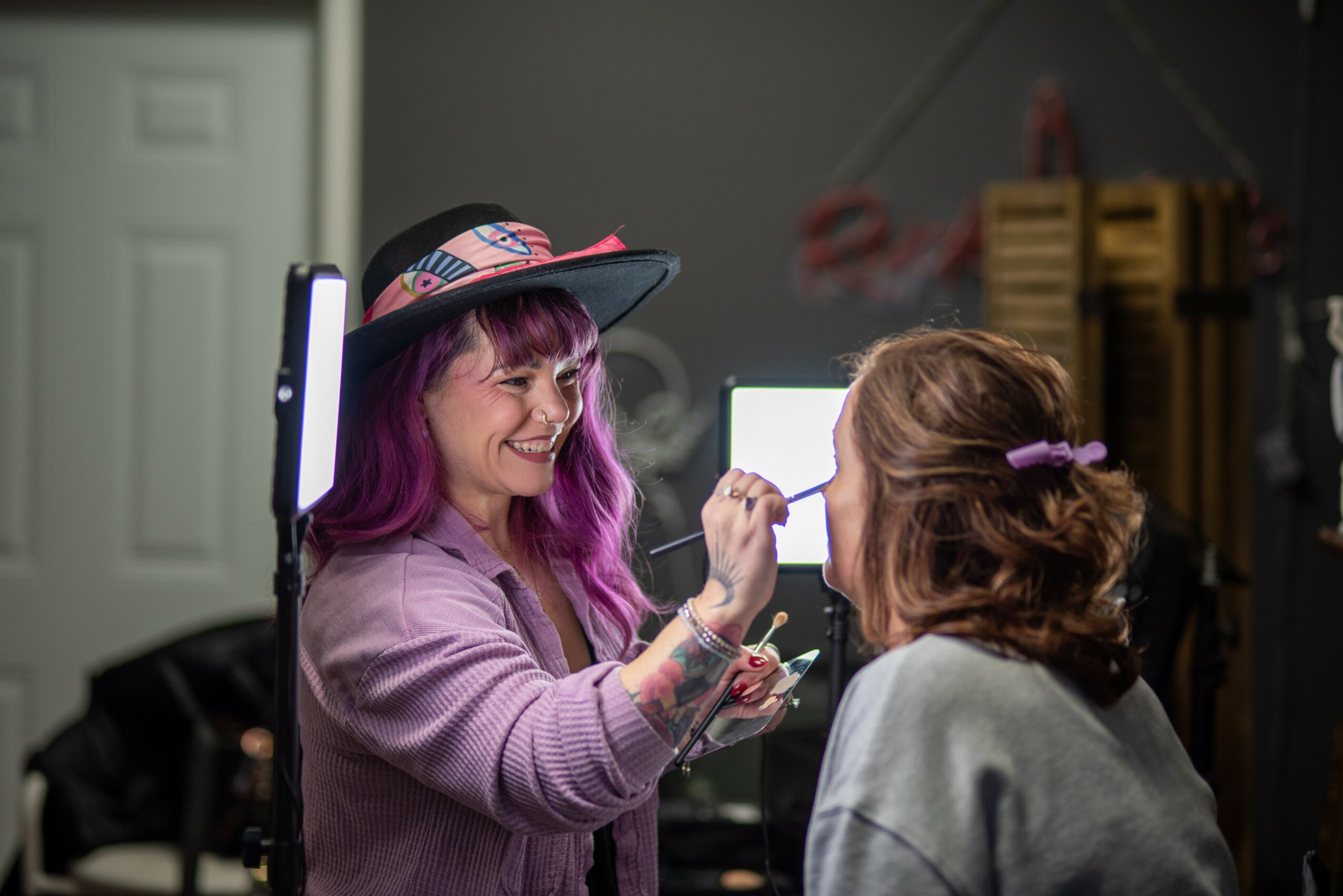 Two women smiling and interacting, one with purple hair and a hat, the other with brown hair, in a workshop setting.