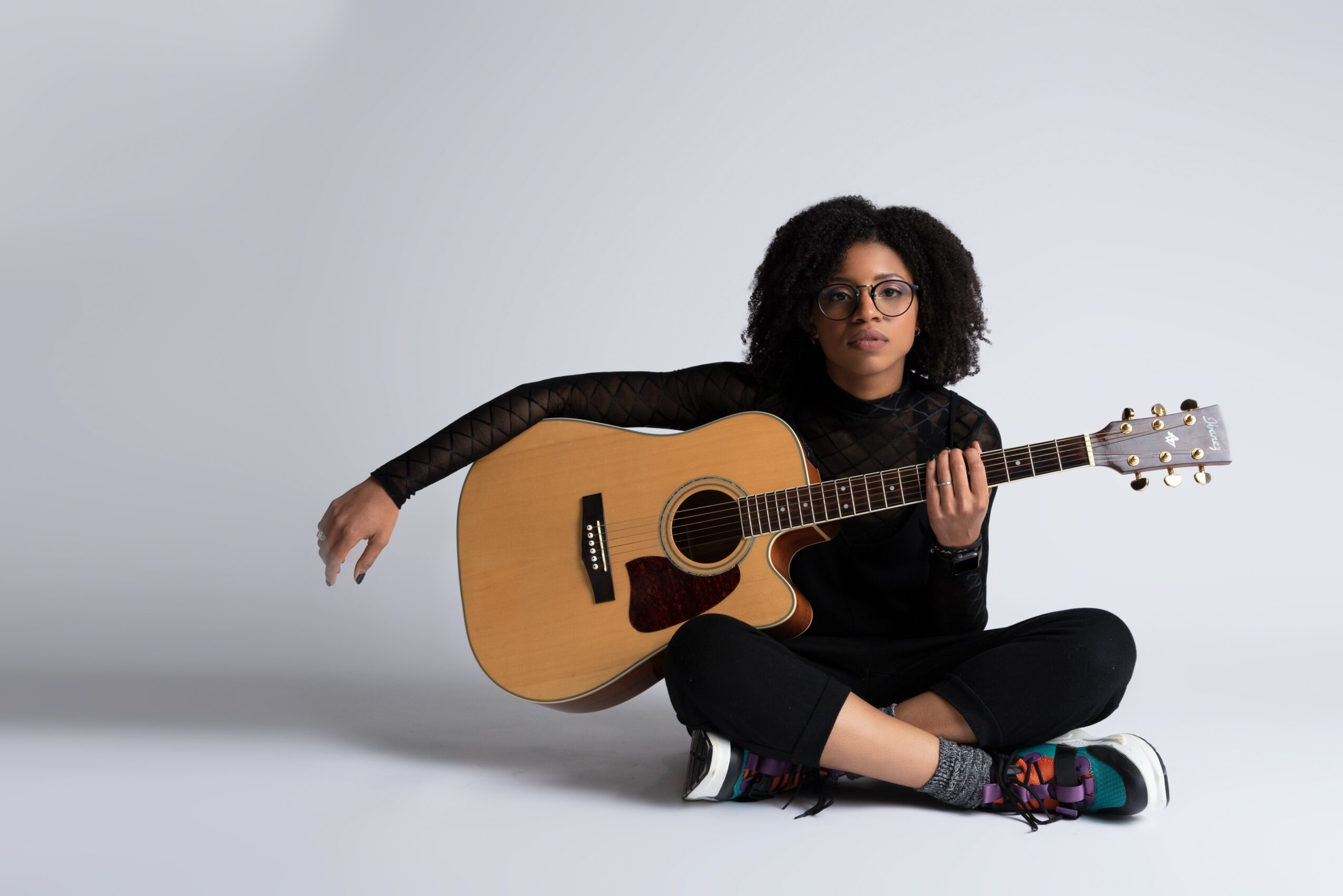 Person sitting cross-legged with an acoustic guitar, wearing glasses and a black outfit, against a plain background.