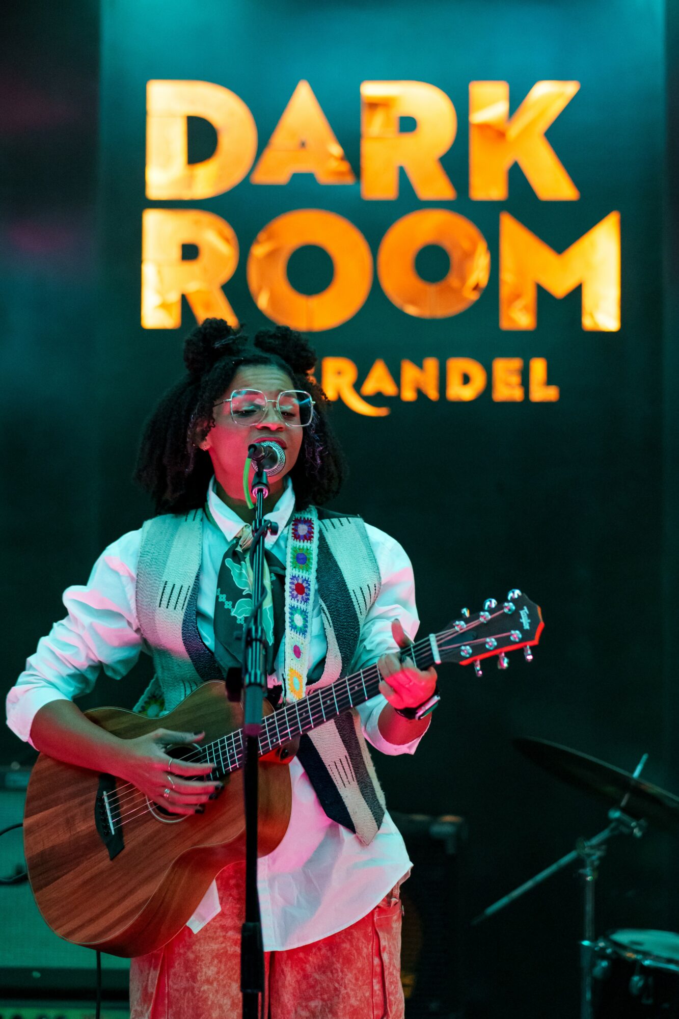 Person playing guitar and singing into microphone on stage with a dark background and illuminated text reading 'DARK ROOM RANDEL'.