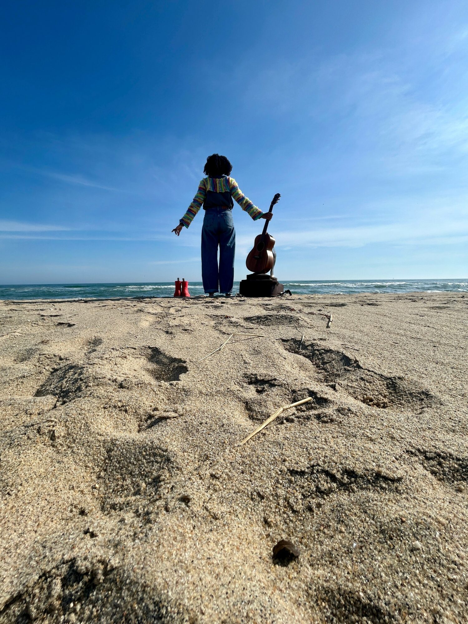 Two people on a sandy beach facing the ocean, one standing and holding a stick, the other sitting, with a clear blue sky.