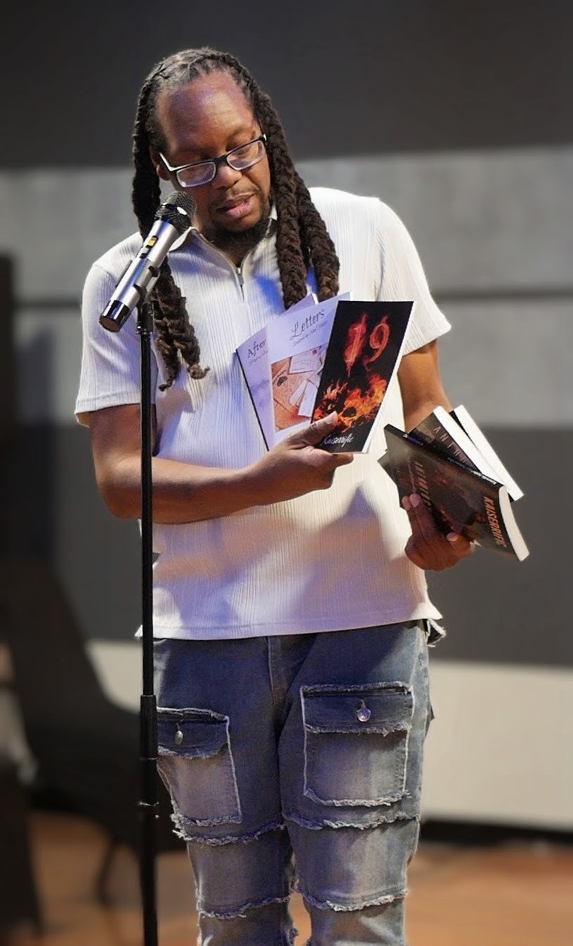 Person with long braided hair and glasses reads from a book at a microphone, holding another book in hand.