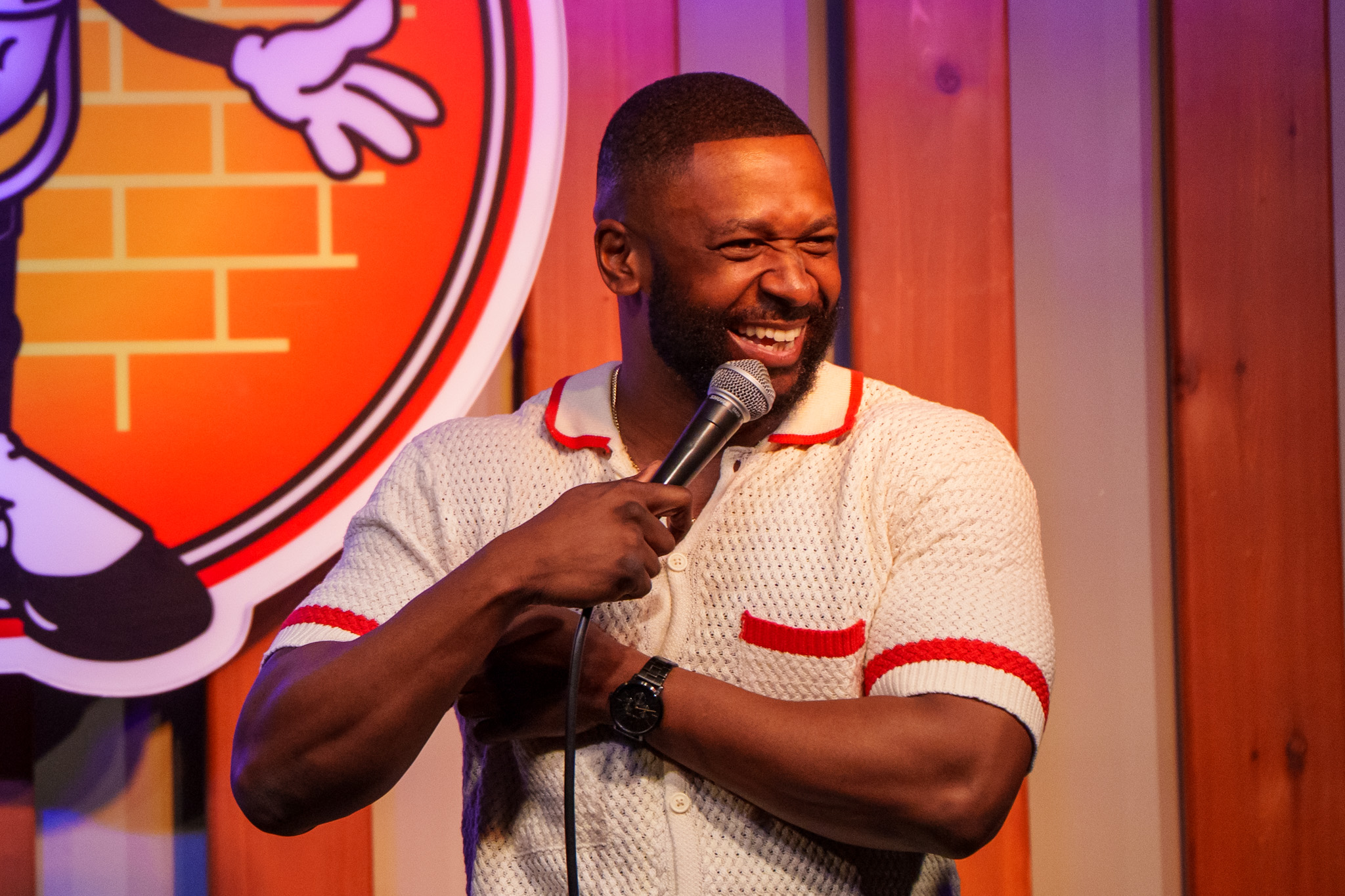 Man laughing and holding a microphone on stage, wooden background, colorful logo partially visible, wearing a light-colored shirt with red accents.