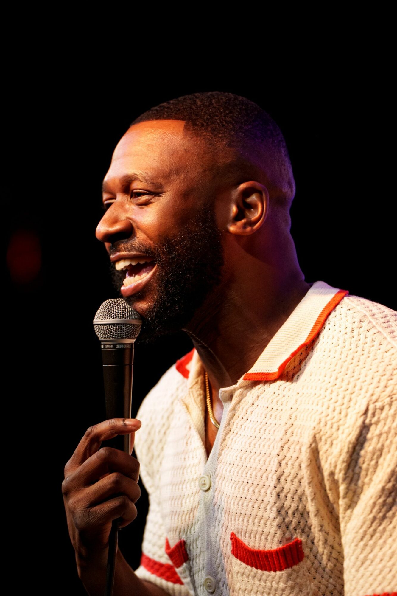 Man speaking into microphone, smiling, with dark background, wearing a light-colored sweater with red accents.