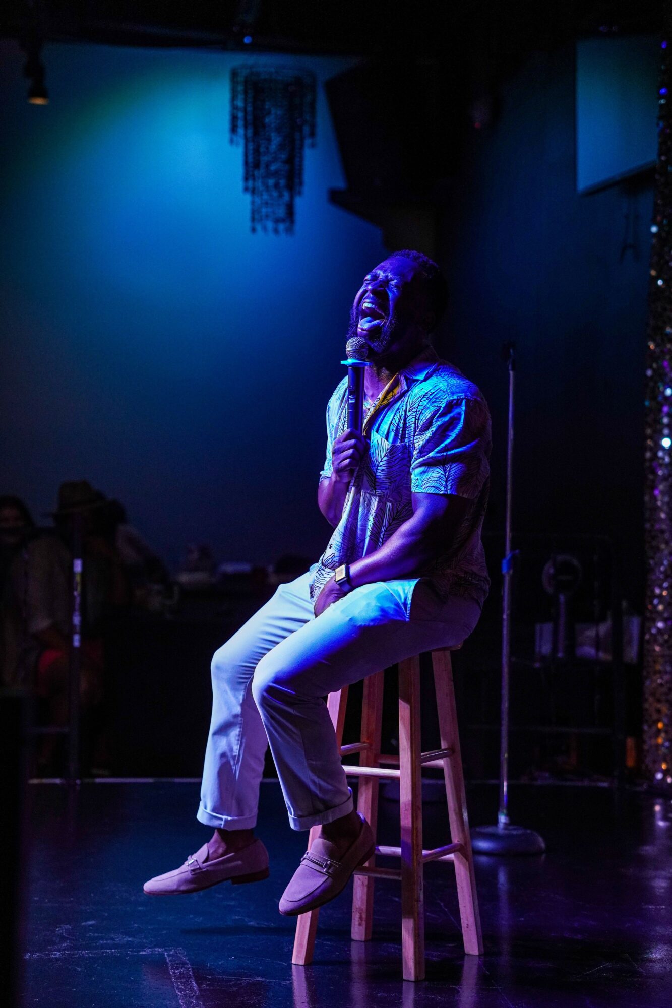 Performer sitting on a stool on stage, illuminated by blue and purple lighting, with a dark background.
