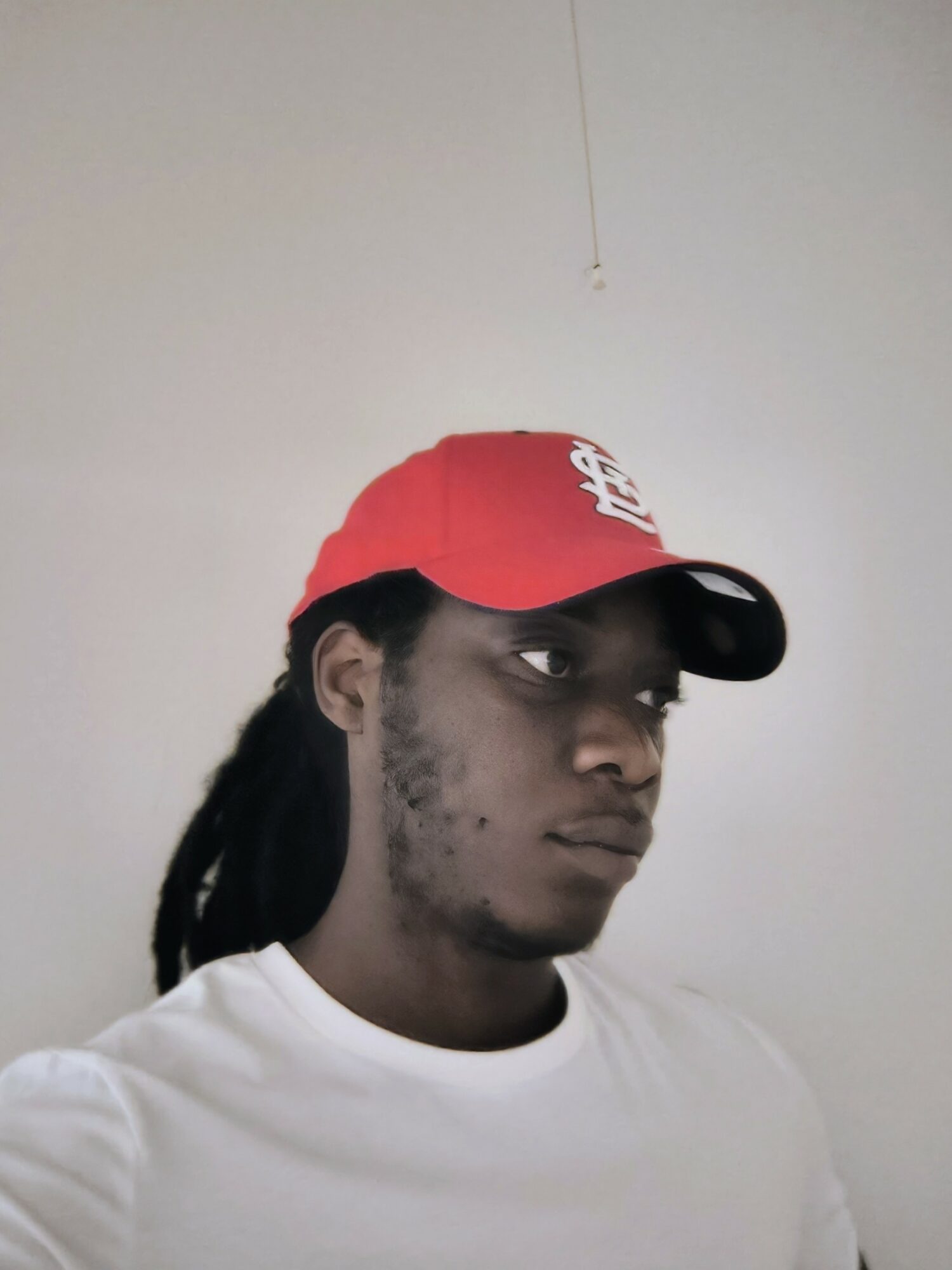 Young man with dreadlocks wearing a red cap and white shirt, looking to the side, plain background.