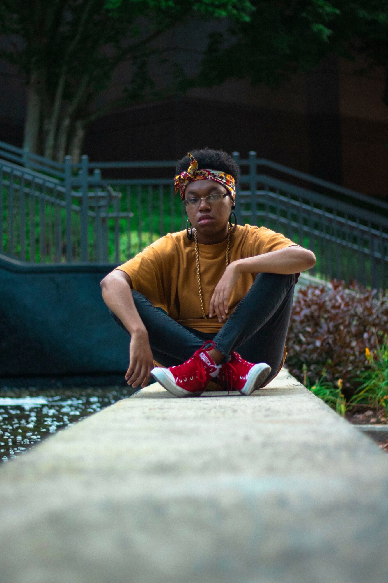Person sitting on a ledge outdoors, wearing glasses, a headband, a yellow shirt, black pants, and red shoes, with greenery in background.