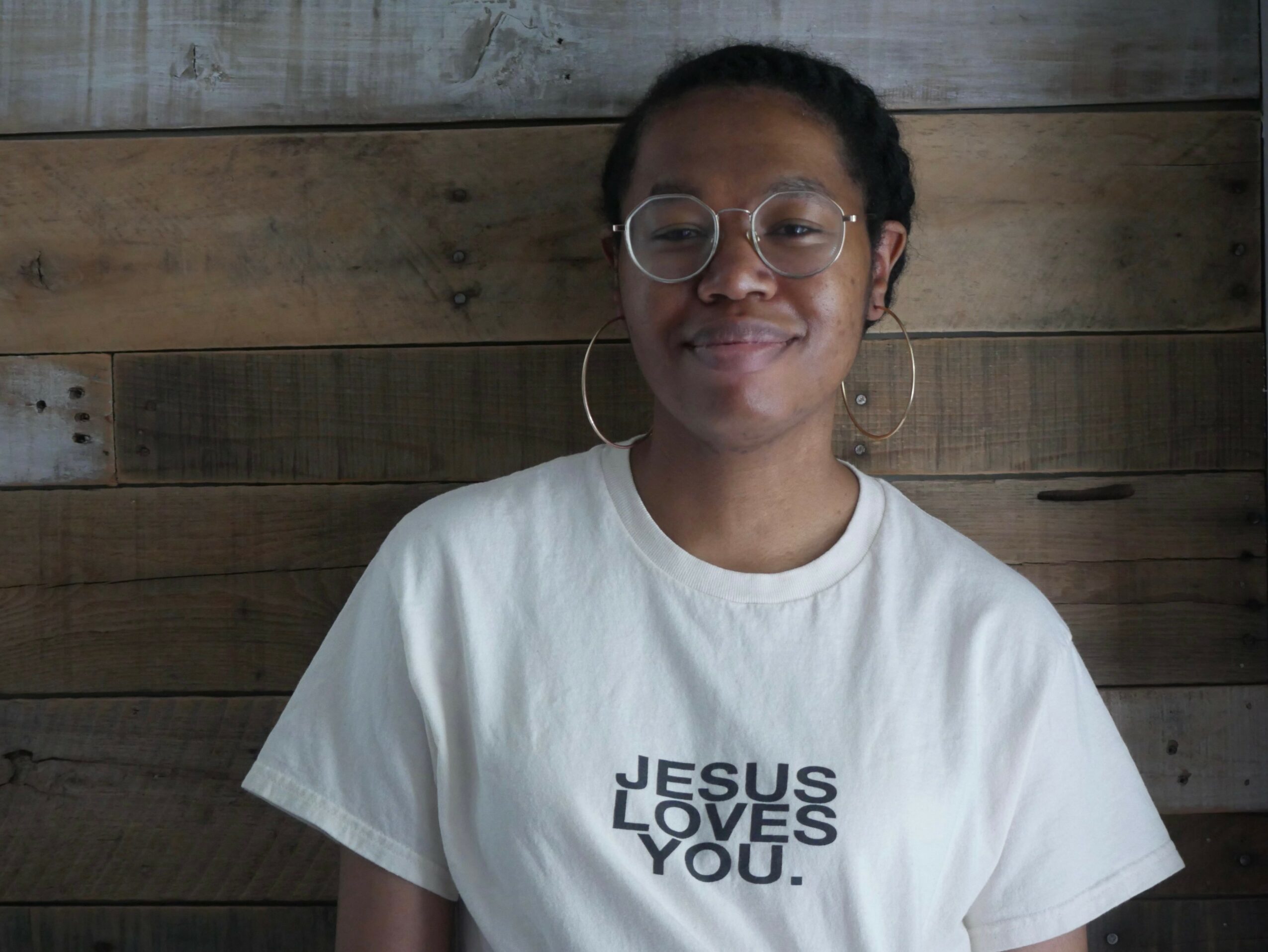 Person with glasses and hoop earrings smiling, wearing a white T-shirt with text, against a wooden background.