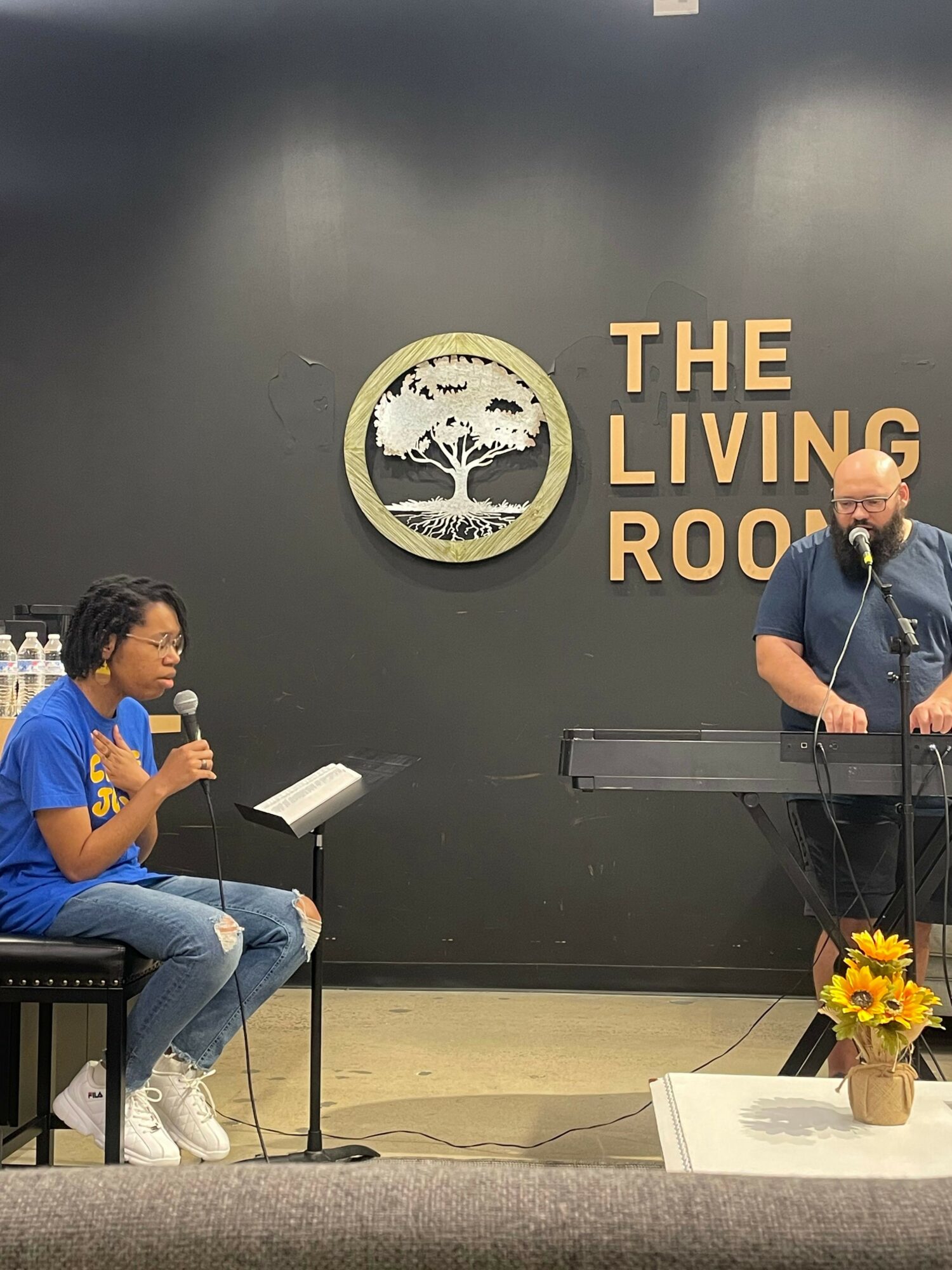 Person speaking at podium with a microphone, seated woman drinking water, black wall with logo reading 'The Living Room', yellow flowers
