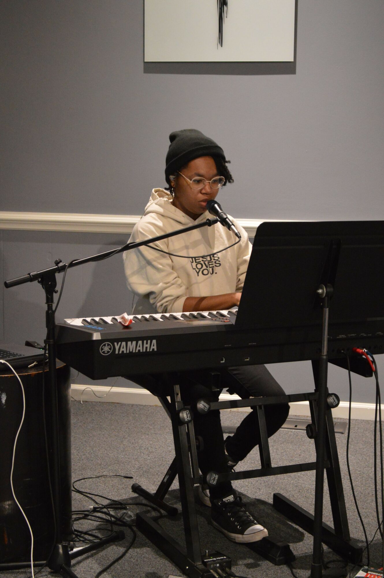 Person playing Yamaha keyboard with microphone in a room, gray wall, and framed picture, wearing glasses and a hat.