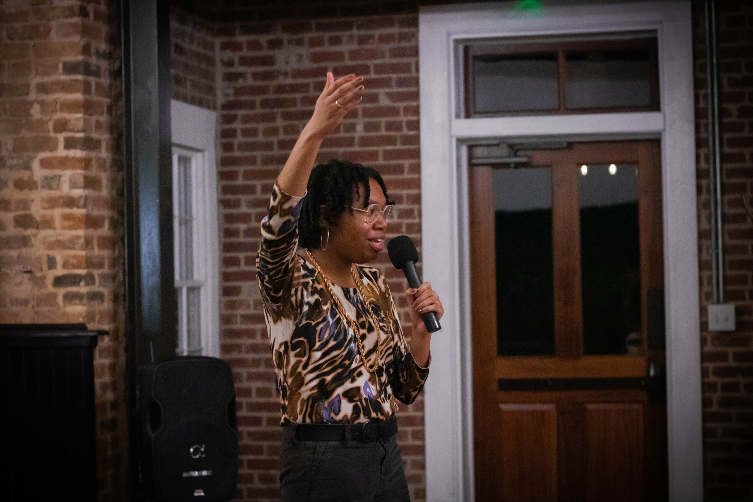 Person with short hair and animal print shirt speaking into a microphone, raising one hand, in front of a brick building.